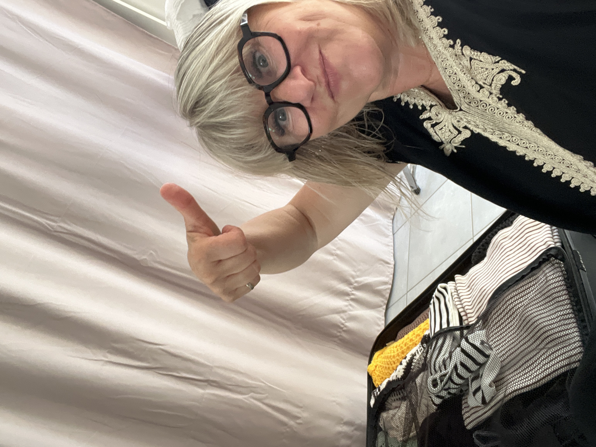 In Chambœuf, France, a woman with blonde hair and glasses is taking a selfie. She is wearing a black garment with ornate gold embroidery around the neckline. Her left arm is raised, and she is giving a thumbs-up gesture. In front of her is an open suitcase filled with neatly folded clothing, including items with stripes and a bright yellow knitted piece. A light-colored curtain hangs in the background. The scene suggests preparation for travel.