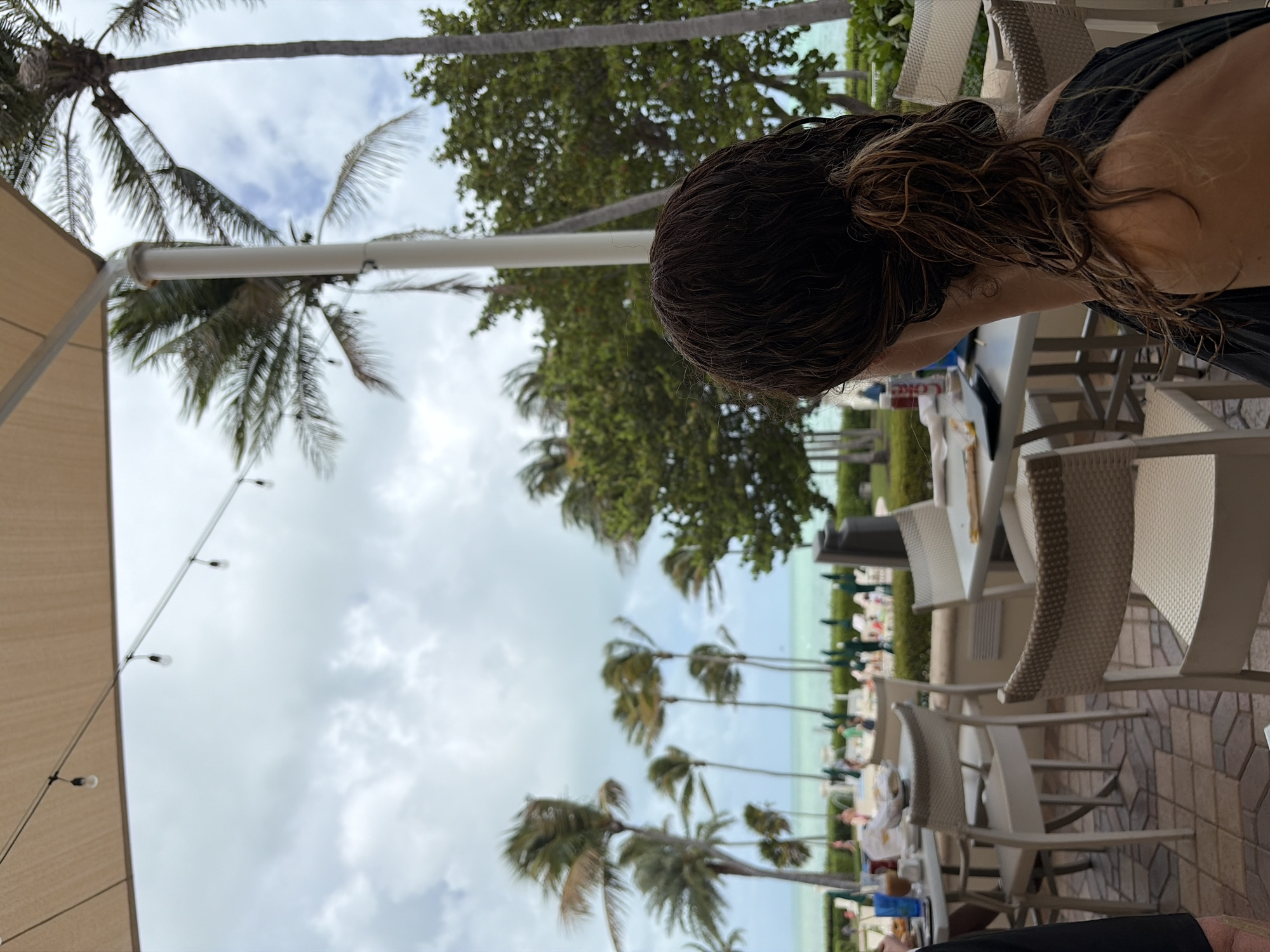 The image shows a view from an outdoor seating area. Palm trees are visible in the background against a cloudy sky. The seating area has white chairs and tables. A person with dark, wet hair is partially visible in the foreground. In the distance, people are gathered near the beach. It is a beachy location on Key Biscayne.