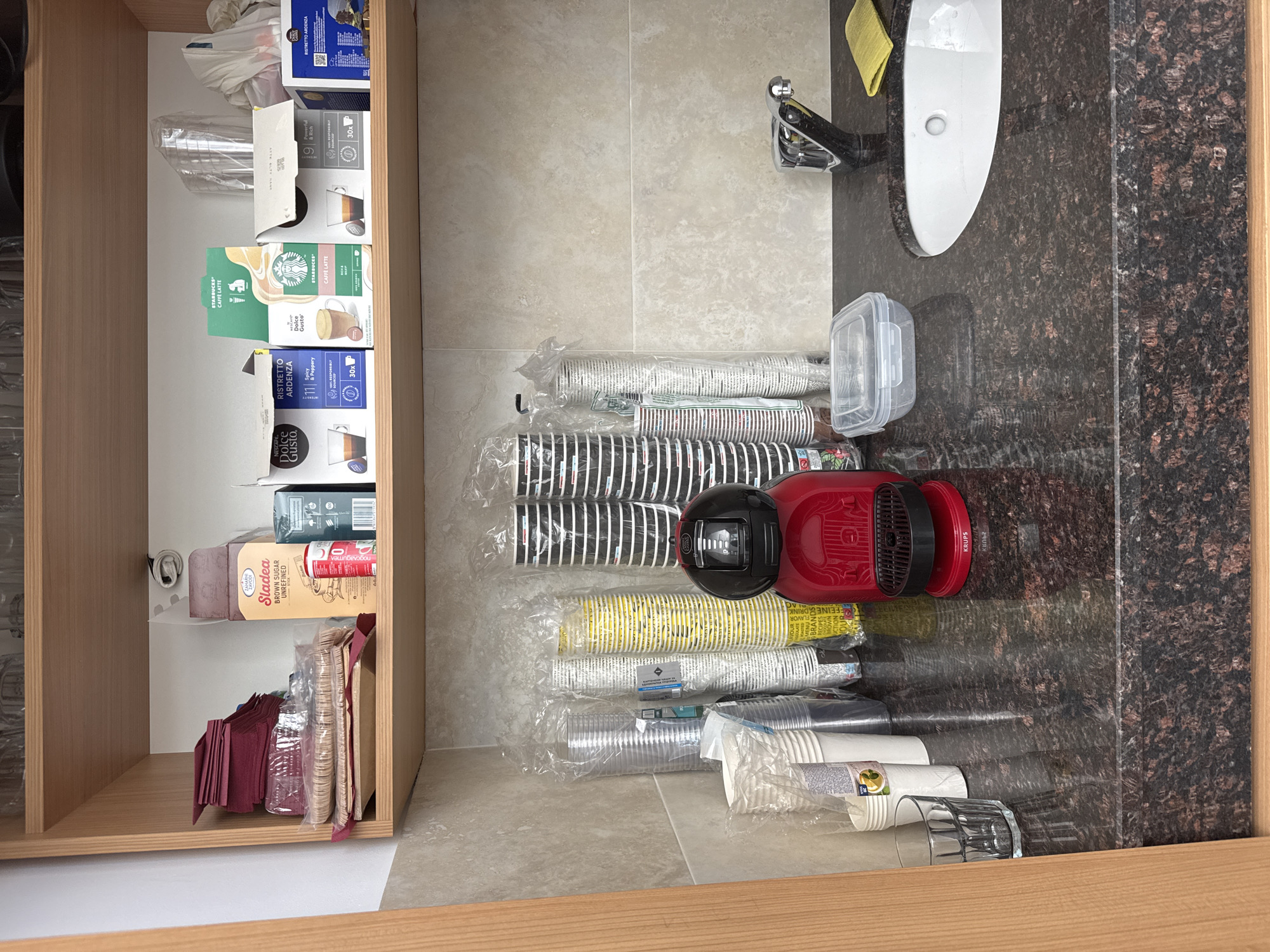 The image shows the inside of a drawer, likely in a kitchen or kitchenette setting. The drawer is filled with various items including coffee capsules from brands like Dolce Gusto and Starbucks, cups, coffee machine, napkins, sugar and some kitchenware. The overall organization of the drawer seems somewhat cluttered. The environment includes a countertop surface with a built-in sink, suggesting the drawer is located in a kitchen or bathroom cabinet. There is no information to pinpoint this scene to Sofia, Bulgaria, or the presence of any people.