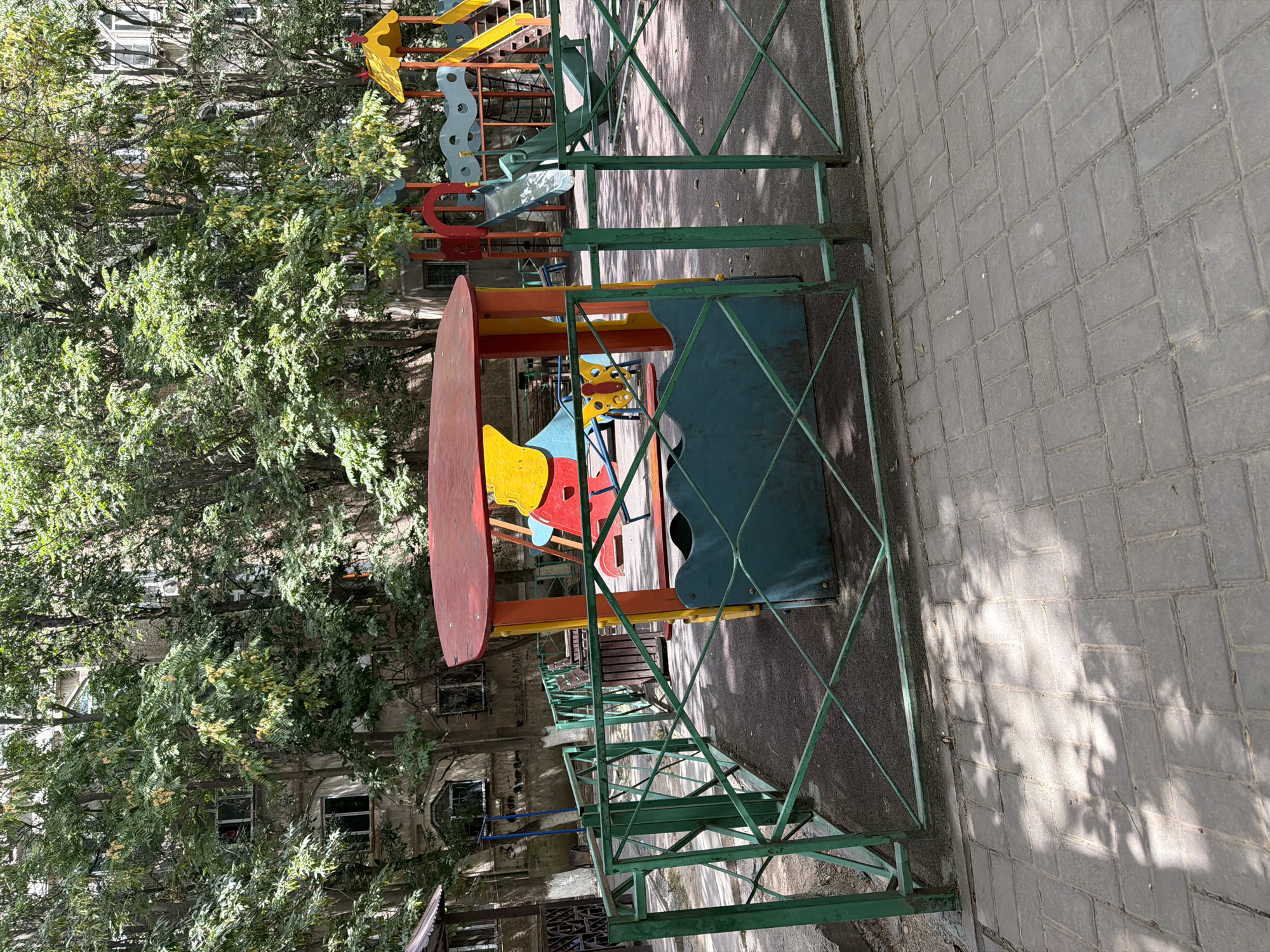 The image shows a children's playground in a city courtyard in Shymkent, Kazakhstan. The playground features a colorful climbing structure with slides and a small playhouse, all enclosed by a green metal fence. Trees with lush green foliage surround the playground, casting dappled shadows on the paved ground. In the background, a multi-story apartment building is visible, with windows peeking through the dense leaves. The pavement in the foreground is made of grey rectangular stones. No people are visible in the image.