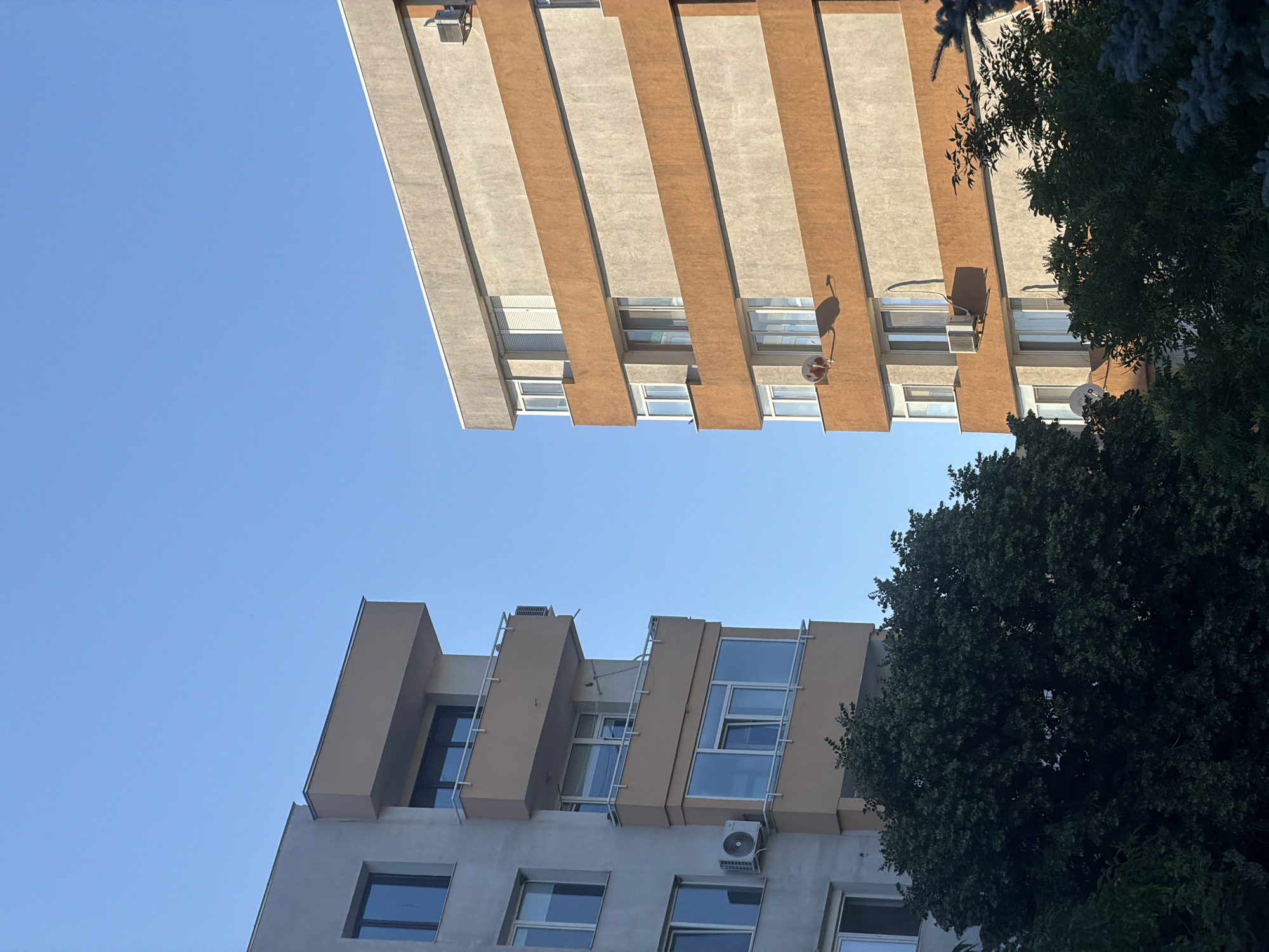 The image presents a low-angle view of two buildings in Blagoevgrad, Bulgaria, taken on a clear day. The sky is bright blue. The building at the top of the frame has a modern facade with alternating beige and gray vertical stripes. Air conditioning units and satellite dishes are mounted on its exterior. The building below it also features a modern design, with beige protrusions framing windows. A tree with dense, dark green foliage partially obscures the right side of both buildings.