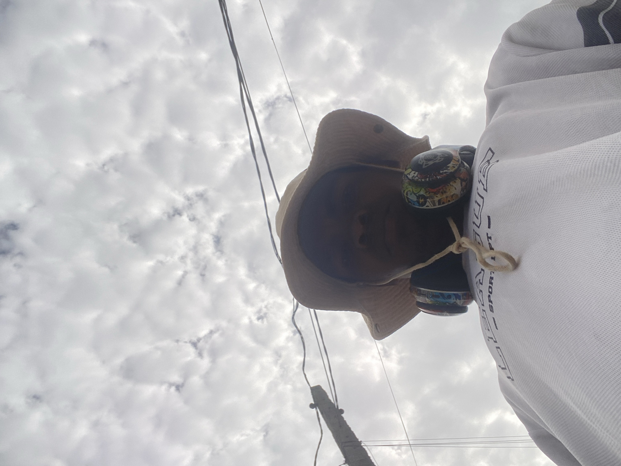 The photograph, taken from a low-angle perspective, features a person wearing a tan hat and headphones against a backdrop of cloudy sky and power lines. The individual is also wearing a white athletic shirt with black lettering. A utility pole is visible near the bottom of the frame. It is difficult to discern identifying markers of Ado, Nigeria within the image.