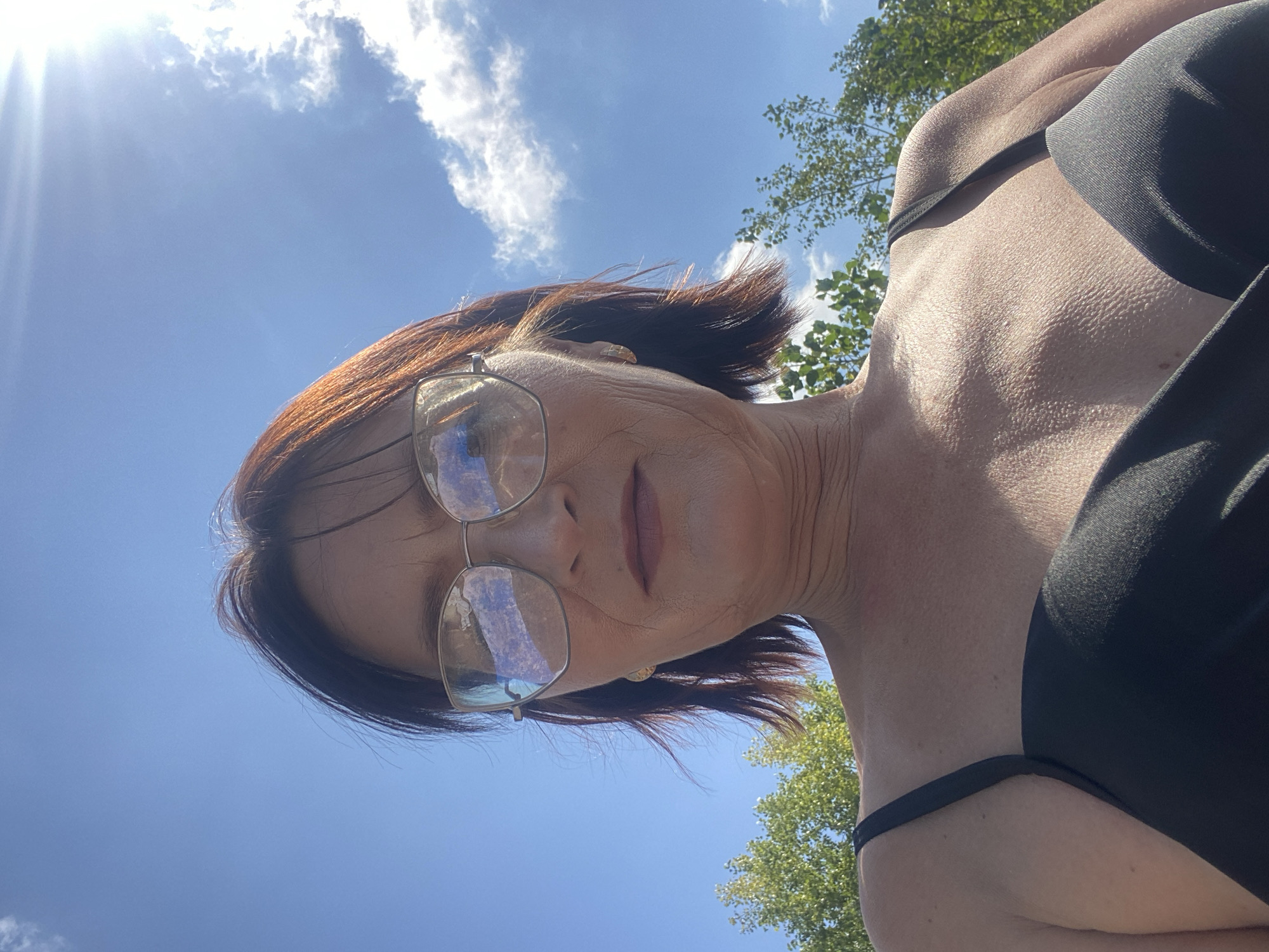 A medium shot, from a low angle, shows a woman with auburn hair and glasses, looking up into a bright blue sky with some clouds. She is wearing a black tank top. In the background, green trees are visible against the sky. The image is framed from the chest up, with the focus on the woman's face and upper body. The scene appears to be outdoors, possibly in a park or natural setting.