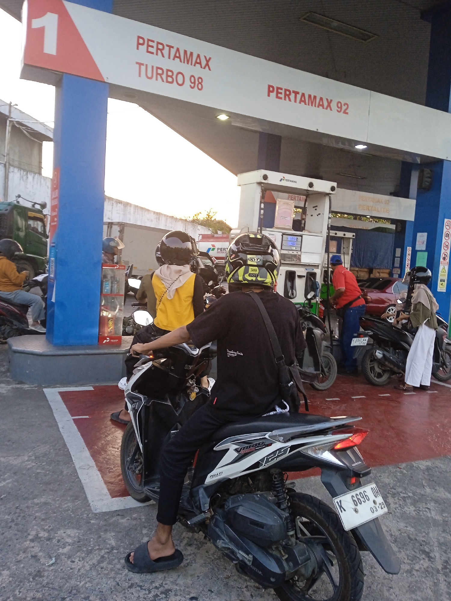 The image depicts a scene at a Pertamina gas station, likely in Semarang, Indonesia. The primary subjects are several individuals on motorcycles, waiting or in the process of refueling. A prominent gas pump is visible, with a person in a red shirt and blue pants operating it. Above the pump, signs indicate fuel types: "PERTAMAX TURBO 98" and "PERTAMAX 92".
In the foreground, a person wearing a black t-shirt and black pants, with a black helmet, is seated on a white and black motorcycle. They are wearing dark gray slip-on sandals. Behind them, another motorcycle has a rider in a yellow vest and a beige headscarf, also wearing a helmet. Further back, another person in a helmet is visible on a motorcycle.
To the left, a green truck is parked near a blue gas pump column. The ground is paved and marked with white and red lines. The overall environment suggests an outdoor, urban setting during daylight, with a warm, possibly sunset or sunrise, light illuminating the scene. The gas station infrastructure, including the pumps and signage, is characteristic of Indonesian gas stations. The motorcycle license plate in the foreground reads "K 6696 OM 03-29".