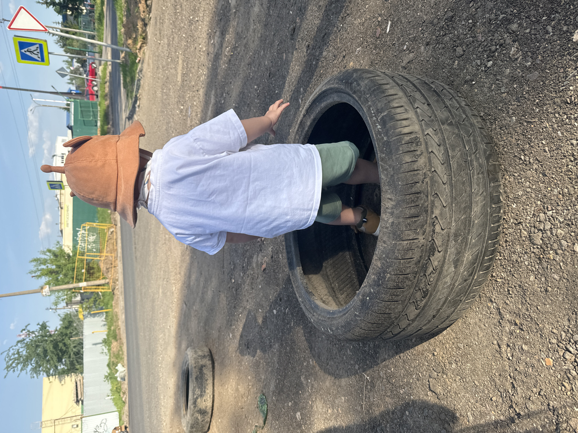 A child in a brown hat is standing inside a car tire on a gravel-covered surface in Akulovo, Gp Kubinka, Russia. The child is wearing a white shirt and green shorts. Another tire is lying on the ground nearby. A road with street signs is visible in the background. A yellow fence is adjacent to the road and behind it is a building. The sky is visible and clear.