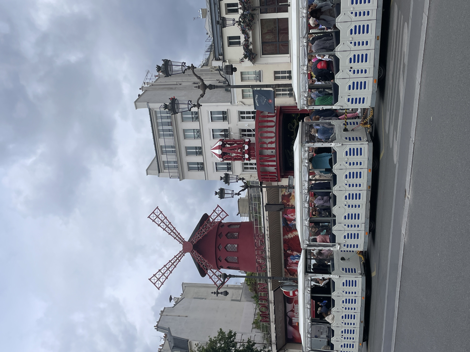This is a portrait-oriented photograph taken in Paris, France, under a cloudy sky. The main subject is the iconic Moulin Rouge, a famous cabaret building with a red windmill on its roof. The Moulin Rouge is painted red and features latticed windmill blades. Next to the Moulin Rouge, a white and blue tourist train is parked on the side of a street. The train is filled with passengers, suggesting a guided tour or transportation service. The street has a pedestrian crossing marked with white lines.
Several buildings are visible in the background, with typical Parisian architecture featuring multiple windows and balconies. The sky is overcast, indicating a potentially cool or rainy day, but the overall lighting suggests it is daytime. There are streetlights with ornate lamp posts on either side of the street.
Visible text includes "MOULIN ROUGE" in large red letters on the side of the building, and "PROMOTRAIN" on a blue flag hanging from a lamppost. Other text is present on posters and signs around the Moulin Rouge, but it is not clearly legible. The scene appears to be a popular tourist spot, with people on the train and in the vicinity of the landmark. The image captures a moment of sightseeing in a well-known Parisian district.