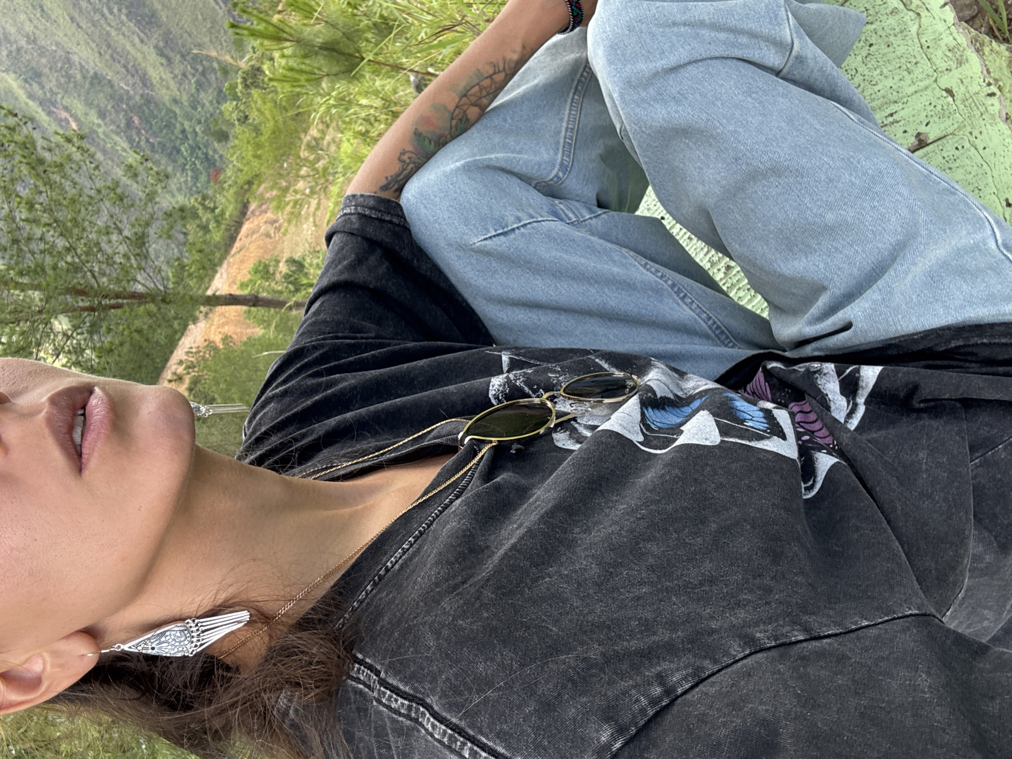 The image shows a person in Lũng Cú Commune, Vietnam, sitting outdoors with crossed legs. They are wearing light blue jeans and a black t-shirt with a graphic print, along with silver earrings and sunglasses. A gold chain necklace is visible. The background features green vegetation and a distant landscape. The person has a tattoo on their forearm. The image is rotated to the left.