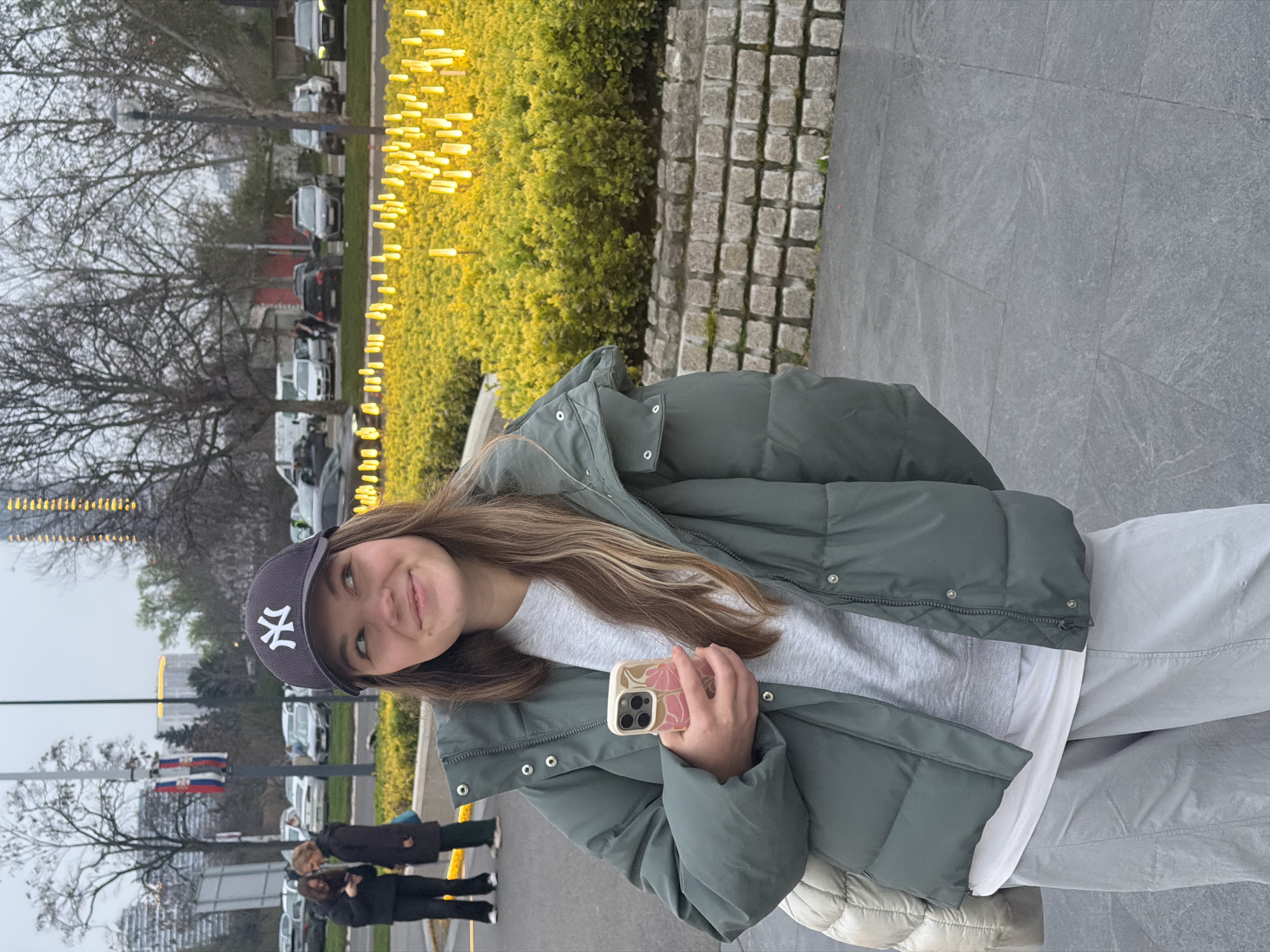 A young woman is standing outside in what appears to be Belgrade, Serbia. She is wearing a dark baseball cap with the New York Yankees logo, a light green puffy jacket, a gray sweatshirt, and light gray pants. She is holding a phone with a floral case. In the background, there are several parked cars, trees without leaves, a row of bright yellow lights, and people walking on the sidewalk. The ground is paved with gray stones. Serbian flags are visible on a pole behind her.