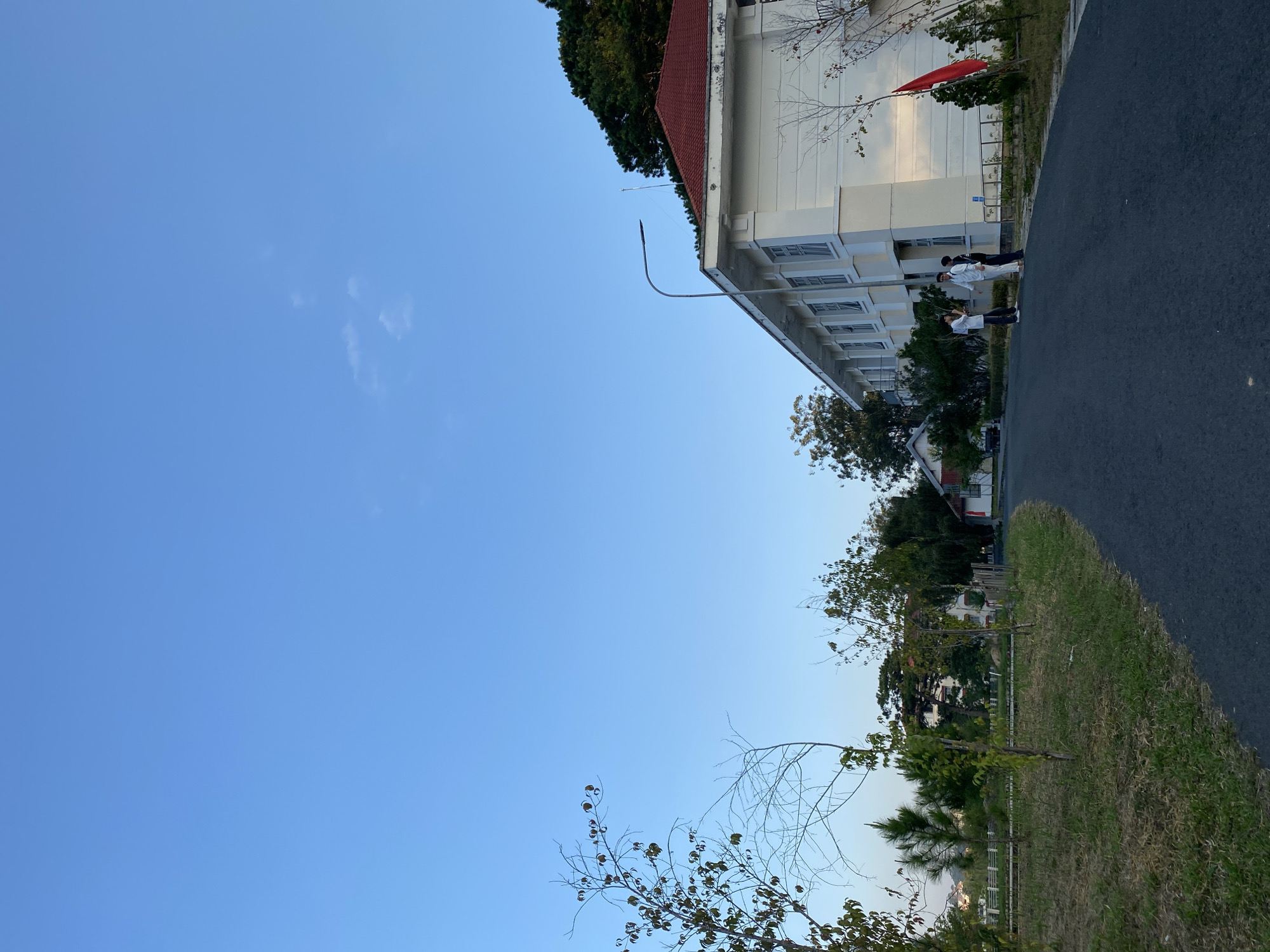 In Lâm Viên - Đà Lạt, Vietnam, a clear blue sky with scattered wispy clouds is visible above a landscape featuring a long, white, multi-story building with a red tiled roof. The building has numerous windows and appears to be an educational or institutional facility. A paved asphalt road curves into the foreground from the right, leading towards the building. On the left side of the road, there is a grassy area with scattered young trees and dry branches. Three individuals, appearing to be young men dressed in light-colored clothing, are standing on the road near the building, engaged in conversation. A bright red flag is affixed to a pole on the side of the building. The environment suggests a temperate climate, possibly late afternoon due to the lighting.