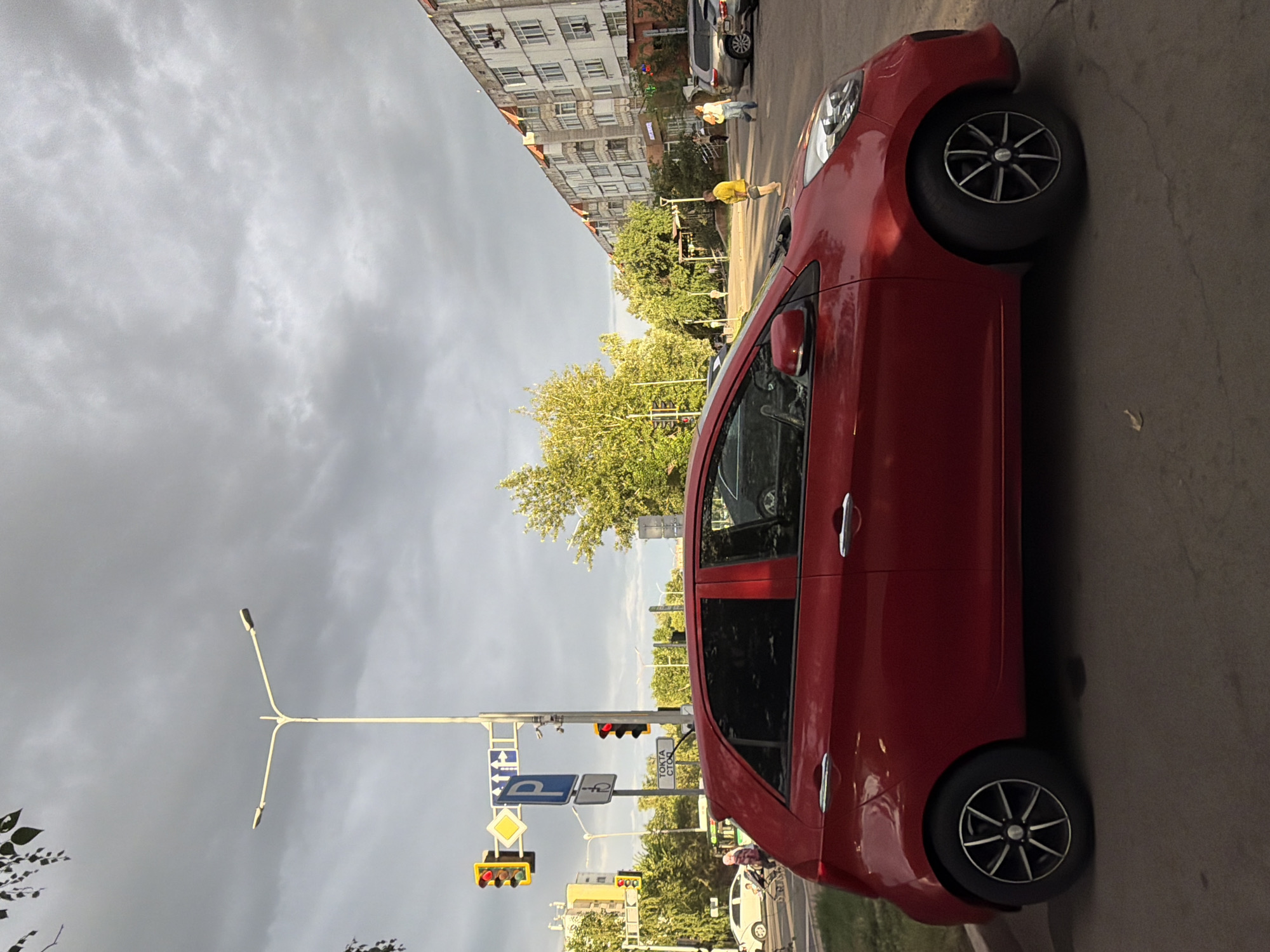 A red hatchback car is parked on the side of a street in Astana, Kazakhstan, under a cloudy sky. In the background, a multi-story residential building with a distinct architectural style lines the street. Across the road, several pedestrians are visible, including a person in a yellow t-shirt and shorts walking across the street, and a woman with a bag. Trees with green foliage line the sidewalks and are interspersed with streetlights and traffic signals. A white car is seen in the distance on the left side of the frame, near a traffic light. The overall scene depicts a typical urban environment in Astana during daytime.