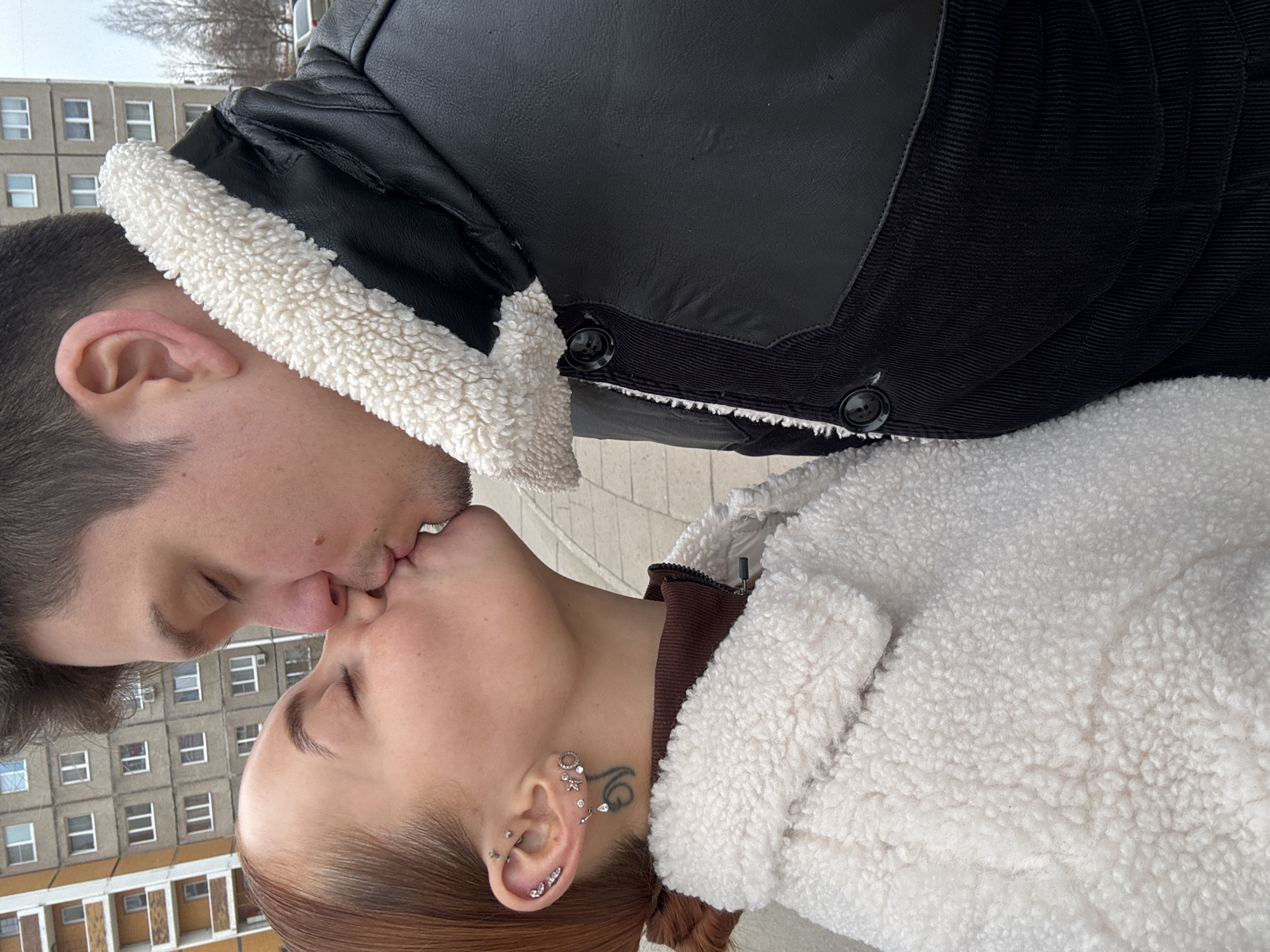 A man and a woman are kissing. The man is wearing a black jacket with a white, fuzzy lining. The woman is wearing a white, fuzzy jacket and has red hair and multiple ear piercings. A building is visible in the background, consisting of windows and yellow balconies. The location of the image is likely Chaikovsky, Russia.
