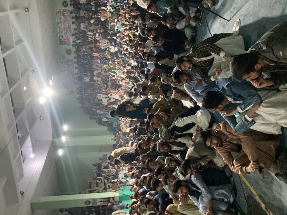 An indoor, wide-angle view depicts a large assembly of several hundred men within a hall in Quetta City Tehsil, Pakistan. The majority of attendees are seated on the light-colored floor, primarily dressed in traditional Shalwar Kameez and various head coverings. Individuals in the foreground are seated cross-legged; one is holding a smartphone horizontally. In the middle ground, several men are standing, including one wearing a dark jacket and another in a light brown jacket near a black camera tripod equipped with recording equipment. The back and left walls are light green, illuminated by overhead fluorescent lights and multiple bright spotlights creating lens flares. Two framed banners with portraits and text are visible on the back wall. Elevated tiered seating areas are present along the perimeter, also occupied by attendees. The crowd is predominantly attentive, facing towards an unseen focal point.