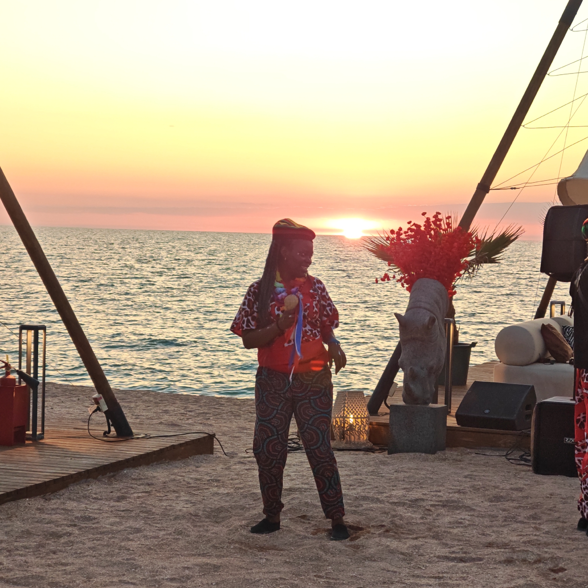 A person stands on a beach during sunset, holding a maraca and wearing a colorful patterned shirt and pants. The person has dreadlocks and a multi-colored hat. In the background, the sun is setting over a calm body of water, casting a warm glow across the sky. A wooden platform with a fire extinguisher and lighting is visible to the left. To the right, there are speakers, lounge furniture, and a sculpture of a rhinoceros head adorned with red flowers and palm fronds. The scene is set in Мысовское сельское поселение, Ukraine.
