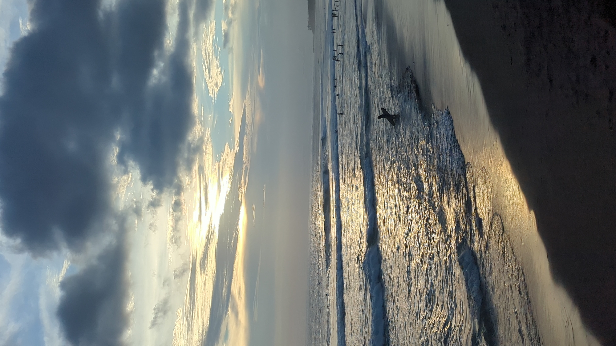 This image captures a dramatic sunset or sunrise scene at a beach in North Kuta, Indonesia, presented in a vertical orientation. The left side of the frame is dominated by the sky, which features a dynamic blend of dark, heavy clouds and brightly lit, golden-hued clouds closer to the center, where the sun's warm glow emanates. Patches of blue sky are visible peeking through the cloud cover.
The right half of the image depicts the ocean and the sandy beach. The ocean's surface shimmers with intense golden reflections from the low-lying sun, as gentle waves roll horizontally across the frame. Numerous small, dark silhouettes of people are visible further out in the water, appearing to be wading or participating in water activities. Closer to the bottom right, a distinct dark silhouette of a person on a surfboard is prominent in the water, indicating surfing.
The sandy beach occupies the far right portion of the image. A large, dark shadow falls across the bottom right section of the sand, creating a strong contrast with the brightly illuminated water and hinting at a nearby object or structure casting a long shadow. The overall atmosphere is serene and vibrant, capturing the picturesque beauty of the golden hour at this coastal location. There is no visible text in the image.