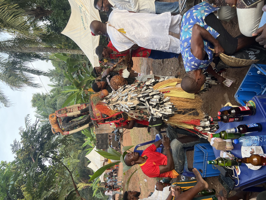 An outdoor cultural gathering in Nnobi, Nigeria, is captured, centered around an elaborately dressed masquerade figure. The masquerade dominates the foreground and midground, adorned with numerous bundles of what appear to be currency notes or paper strips, creating a textured, almost feathery covering. Its costume also features raffia or straw elements, red and orange fabrics, and an intricate, dark headpiece.
Many people are gathered around, observing the masquerade. In the immediate foreground, a man in a red t-shirt sits on a blue plastic chair, looking towards the activity. Nearby, a blue plastic crate serves as a makeshift table, laden with various beverage bottles (some alcoholic, some water) and stacks of money. Another man, wearing a blue floral shirt, bends over, handling money near a wicker basket. Other individuals in casual and traditional attire are visible standing and sitting throughout the scene.
In the background, lush tropical vegetation, including palm trees and banana plants, indicates a verdant, likely rural or semi-rural setting. White canopy tents are erected, providing shade. One tent clearly displays the text "DESIGNER FEES SERVICES". Speakers for a sound system are also visible in the mid-distance, suggesting an organized event. The ground is dirt. The time appears to be daytime under an overcast or bright sky, creating a vibrant yet diffused light. The overall atmosphere is that of a lively community celebration or traditional festival.