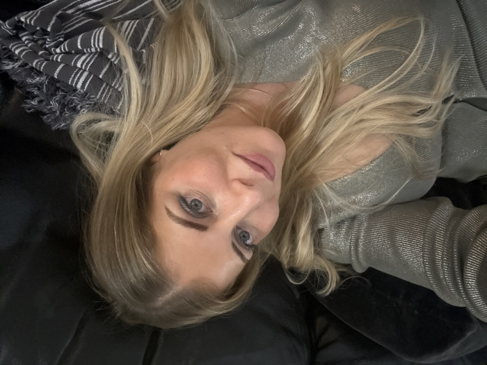 A woman with blonde hair lies on a black surface, possibly a leather couch. She's angled, her face central, gazing upwards with light blue eyes. She wears a silver, shimmery top. To the left, a grey and white striped scarf rests. The lighting is soft, casting gentle shadows across her face.