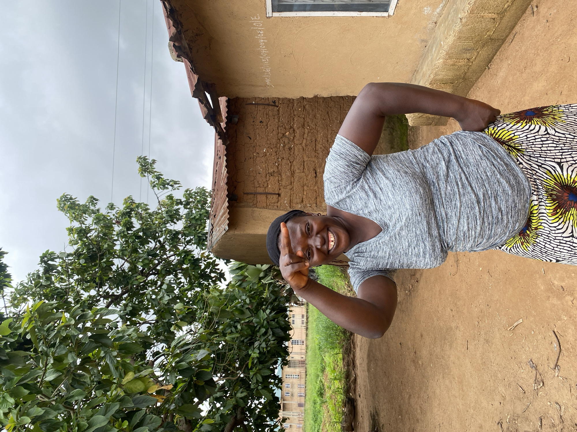 In the city of Riyom, Nigeria, a woman with dark skin and a black headscarf stands outdoors. She is smiling broadly and holding her right hand up to her forehead, creating a gesture. She wears a grey, V-neck t-shirt and a vibrant, patterned skirt featuring yellow, white, and black designs with blue and yellow circular elements. Behind her, a brown clay building with a corrugated metal roof is visible, showing some signs of wear and tear, with a window on the right side. White chalk writing is inscribed on the building's wall. To the left of the building, a large green tree with dense foliage shades the scene. In the background, a long, light-colored building with multiple windows and a tiled roof can be seen, suggesting a more institutional structure. The ground is composed of reddish-brown dirt, scattered with small twigs and dry leaves. The sky overhead is overcast and grey.