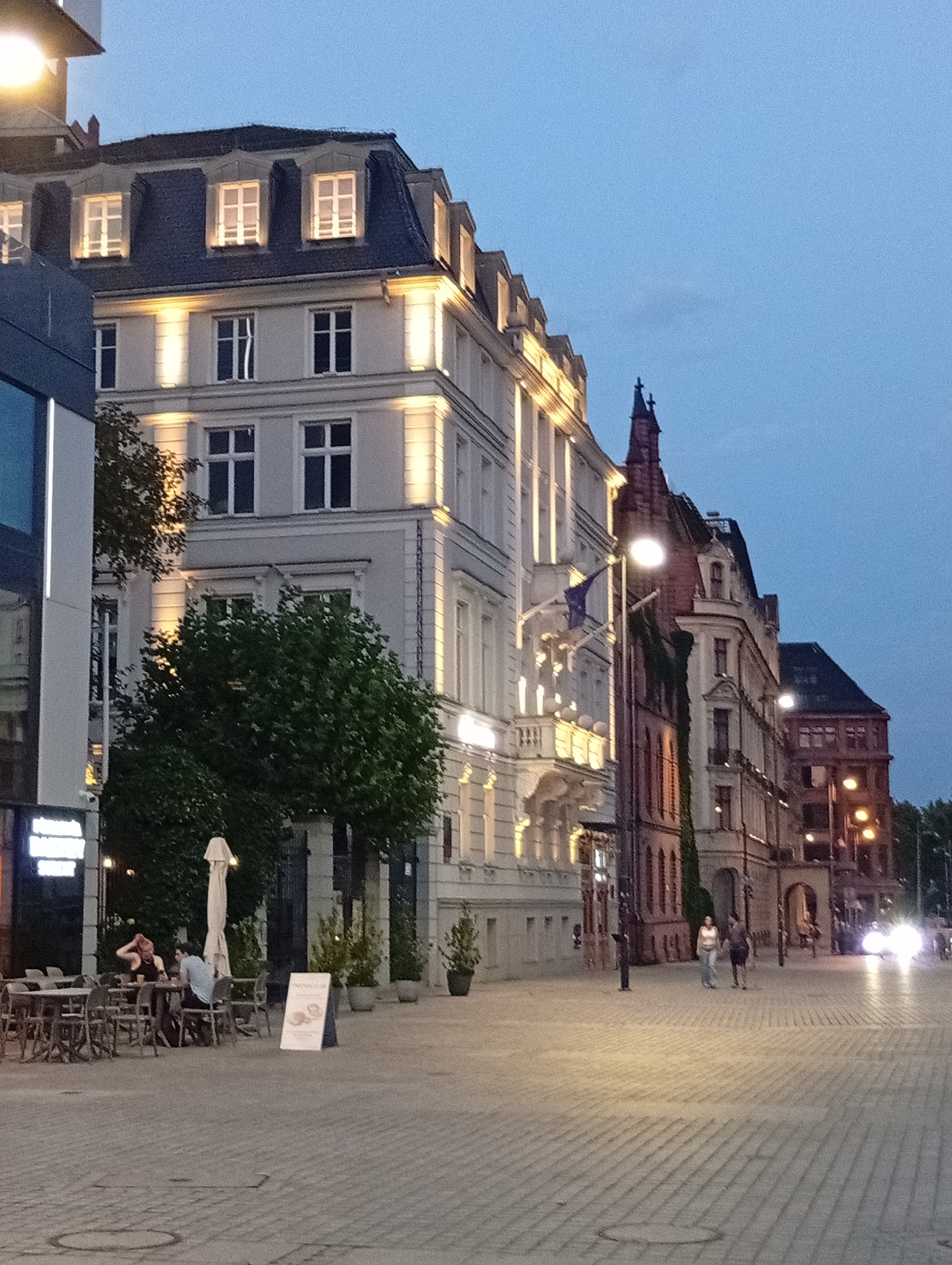 The image depicts a street scene in Wrocław, Poland, at dusk. On the left, a modern building with a glass facade is visible. Adjacent to it is a historic building with a mansard roof, illuminated windows, and decorative lighting. Further down the street, more historic buildings with elaborate architecture are lined up, some with illuminated windows and architectural details.
In the foreground, a cobblestone street leads away from the viewer. Several outdoor cafe tables and chairs are set up on the sidewalk. Two individuals are seated at one of the tables. A white umbrella shades another table. A sign is placed near the tables. Further down the street, two people are walking. The sky is a deep blue, transitioning into dusk. Streetlights are visible, and car headlights are illuminated in the distance. Trees are planted along the sidewalk.