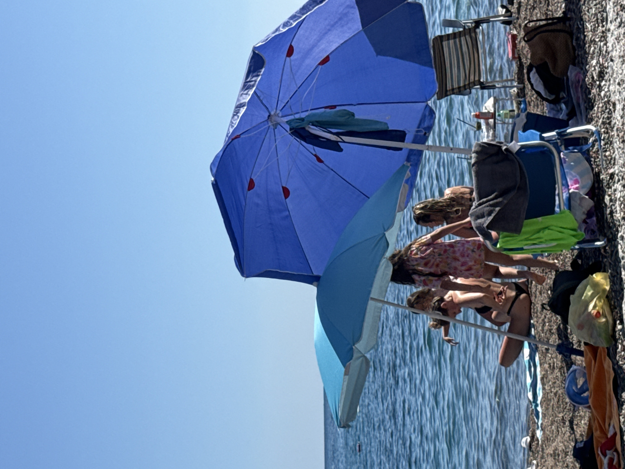 The image captures a sunny beach scene in Rafailovići, Montenegro, under a clear, cloudless blue sky that occupies the upper third of the frame. The horizon line is visible and flat. The sea, a deep blue, is calm with minor ripples extending to the pebble beach in the foreground. A small, dark, rectangular object floats on the water in the mid-distance on the left.
Two open beach umbrellas are prominent. A smaller, light blue umbrella with a white pole stands on the left. To its right, a larger, dark blue umbrella with a white pole features red accents on its underside ribs, and a dark blue and a light blue/teal garment are hanging from its pole.
Several individuals are present on the beach. Partially visible under the light blue umbrella on the left is a person with long, light-colored hair, seemingly sitting or kneeling near the water. To their right, a person with light brown hair tied back is seated or kneeling on the pebbles, facing away from the viewer, wearing a dark bikini bottom. A young person, likely a child, with long light brown hair and wearing a light-colored floral or patterned dress, stands upright with their back mostly to the camera, positioned between the two umbrellas. Another individual is partially obscured by the large blue umbrella on the far right.
Various beach equipment is scattered on the dark pebble beach. These include an orange towel, a dark blue cap, a blue plastic item (possibly a snorkel mask), a translucent yellow plastic bag containing an orange object, and a black backpack in the immediate foreground. A blue foldable beach chair with a grey towel draped over it is visible under the large blue umbrella. On the far right, a striped beach chair (white and dark stripes) with a metal frame is set up facing the water. A small, light-colored table with a red bottle and another container rests near the water's edge behind the chairs on the right.