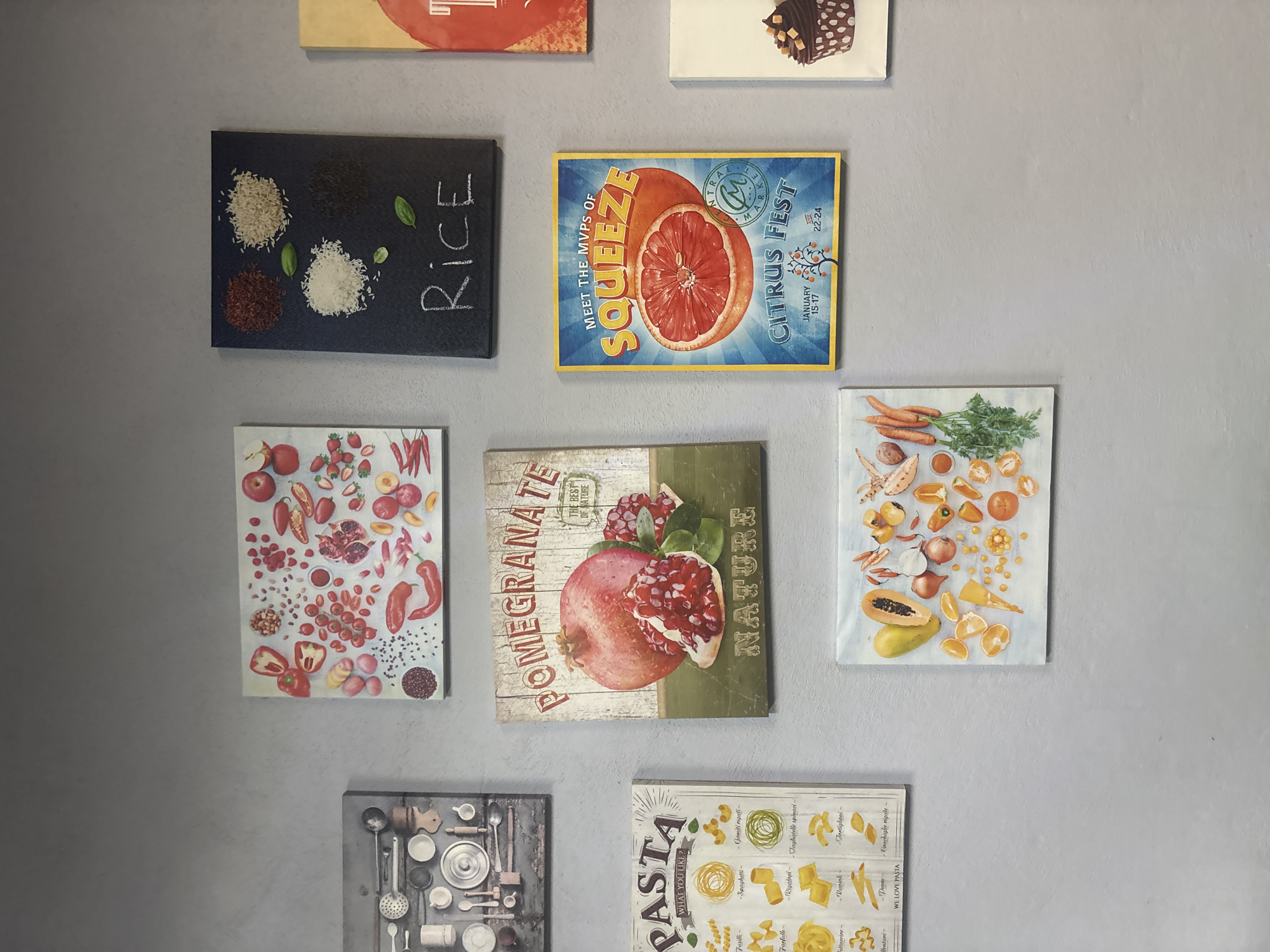 The image shows a collection of food-themed prints displayed on a light grey wall. The prints include images of rice, citrus fruits, pomegranates, an assortment of vegetables, and pasta, each with accompanying text descriptions or related words like "squeeze", "nature", and "what you like?" The image also features a kitchen utensil collage.