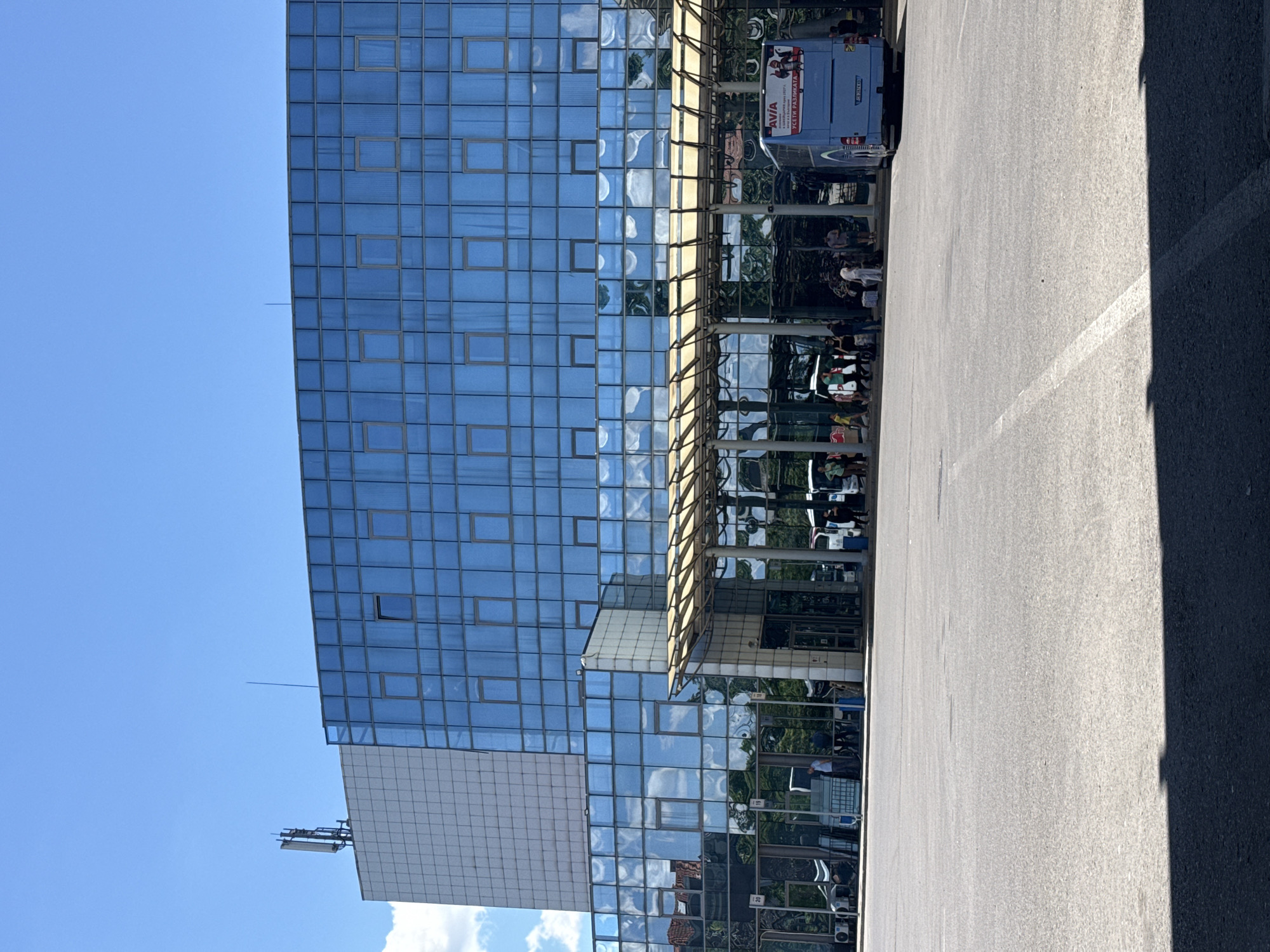 This image depicts the Sofia Central Bus Station in Sofia, Bulgaria. The scene is dominated by the modern, multi-story building of the bus station, characterized by its extensive blue-tinted glass facade and a white, grid-patterned section on the left. A large, open asphalt area, likely a bus parking lot or waiting area, occupies the foreground.
Several buses are visible, including a prominent blue bus on the right side with an advertisement on its rear. Passengers are seen gathered under a covered walkway in front of the bus station entrance, some with luggage. The covered area features a light-colored, sloped roof supported by pillars. People are standing, walking, and interacting in small groups.
The environment is bright and sunny, with a clear blue sky and scattered white clouds. A cell tower is visible on the rooftop of the white section of the building. The reflections in the glass facade show sky, clouds, and surrounding trees. The overall impression is that of a functional transportation hub on a clear day.