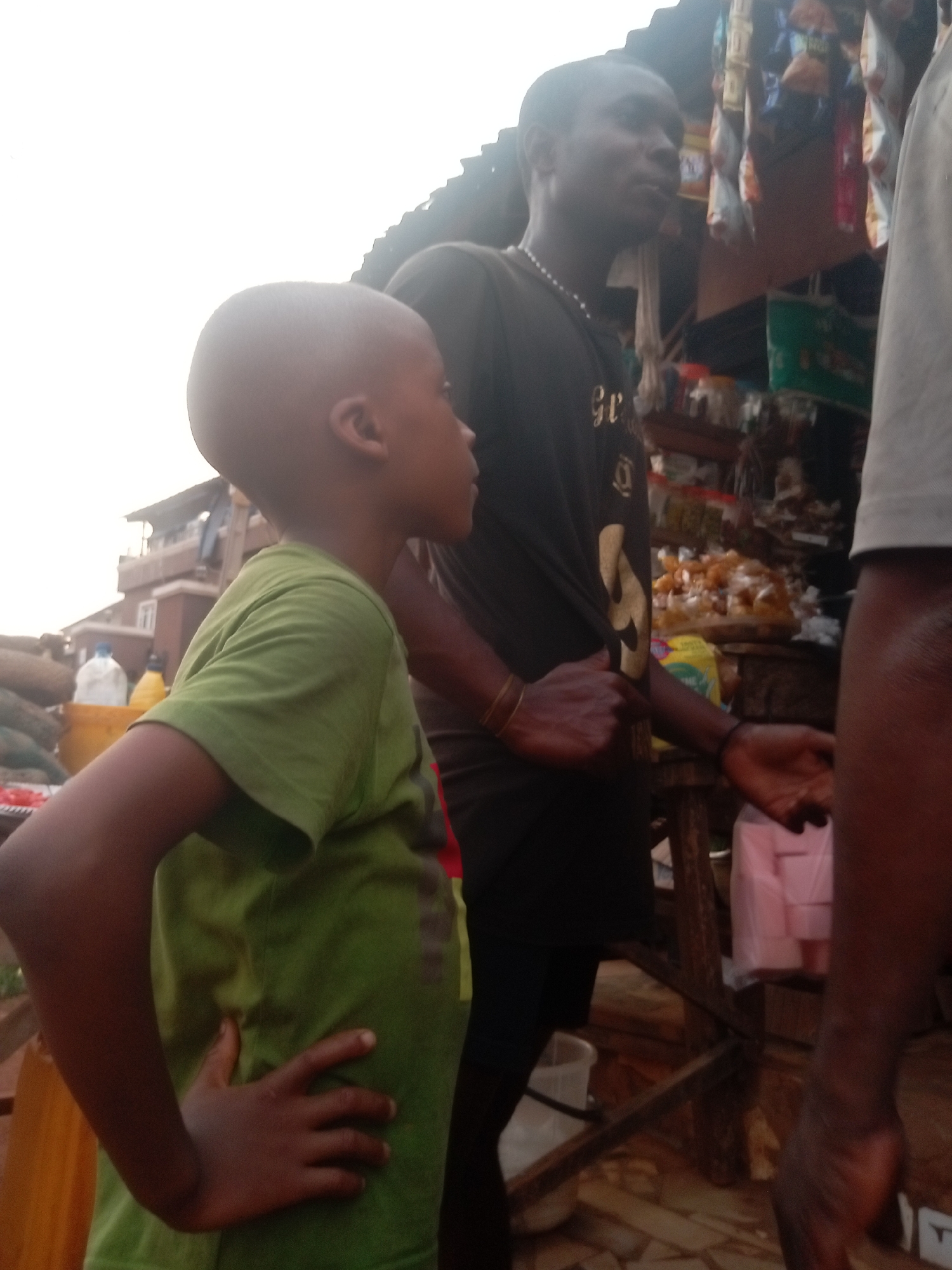 In Ogidi, Nigeria, a young boy with a shaved head stands in a green t-shirt, his hand on his hip, looking to the side. Behind him, a taller man with a black t-shirt and necklace stands with his hand extended, appearing to be offering or asking for something. They are in front of a wooden structure with various items displayed, including snacks and packages, suggesting a market or street vending setup. Another individual is partially visible on the right edge of the frame, obscuring the view. In the background, buildings can be seen, indicating an urban environment.