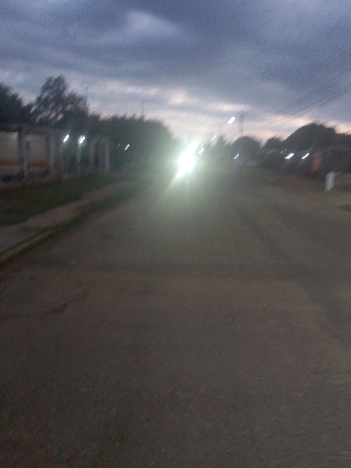 An asphalt road stretches into the distance, illuminated by bright headlights approaching. A building lines the left side, intermittently lit with street lamps. Trees are visible beyond the building, reaching towards a cloudy sky. On the right, darker trees stand near another building, partially obscured by the glare. The overcast sky suggests either dawn or dusk.