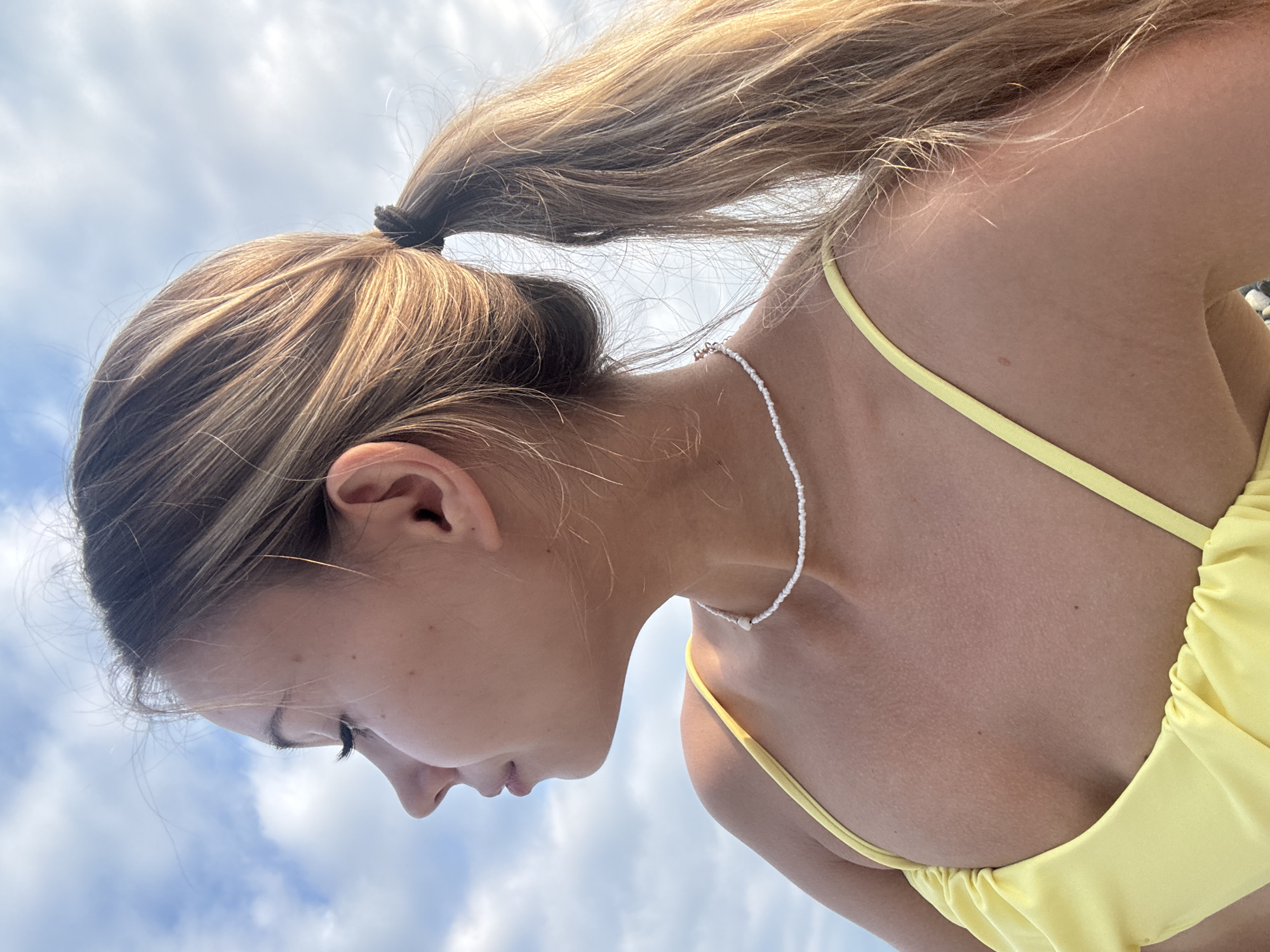 A young woman with blonde hair pulled back into a ponytail is shown in profile, looking to the left. She is wearing a yellow bikini top and a white beaded necklace. The background is a cloudy sky. The image was taken outdoors in Sokhumi, Abkhazia.