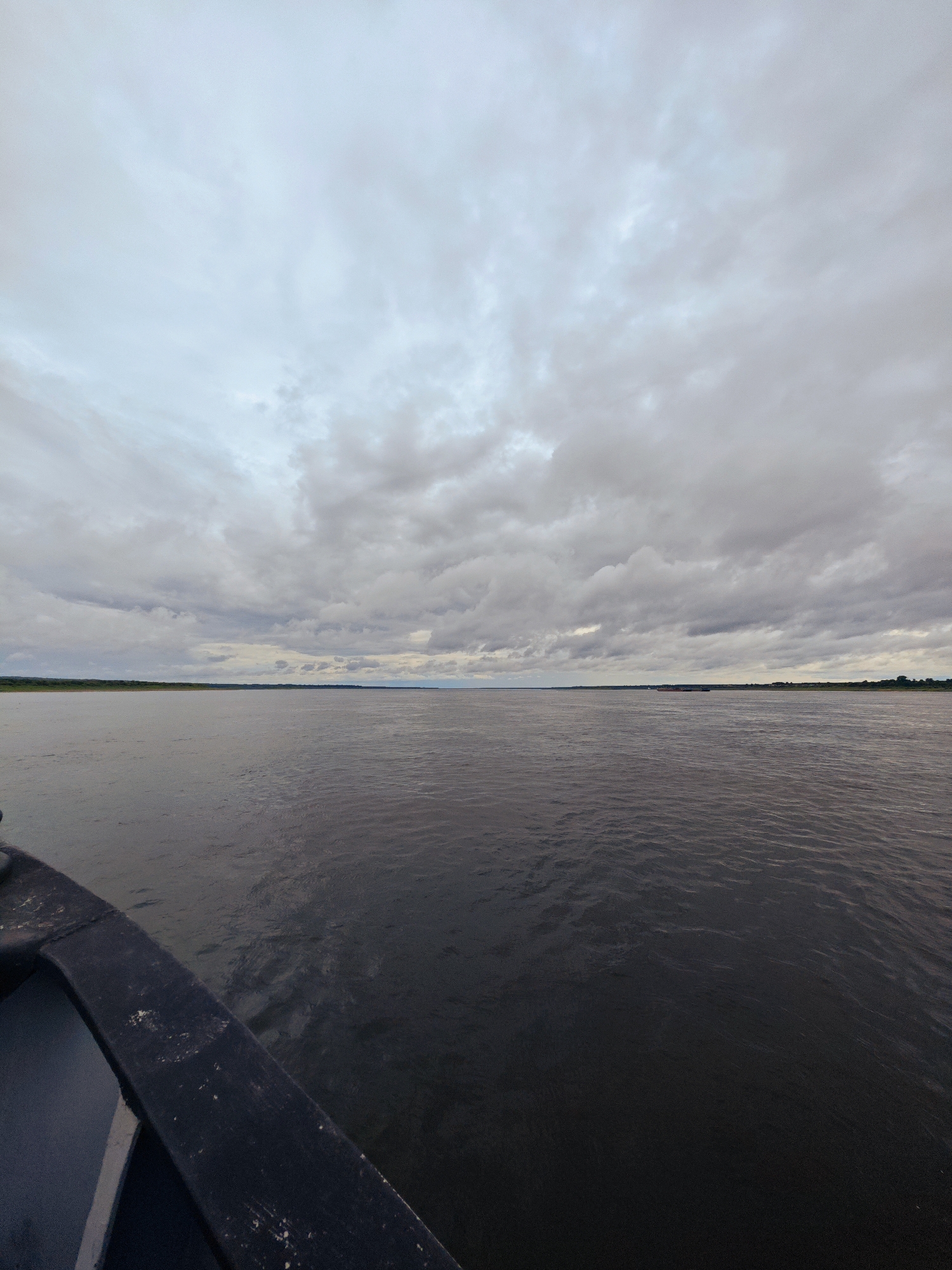The image depicts a wide, calm river under a cloudy sky. The perspective is from the edge of a dark-colored boat, with a portion of its railing visible in the foreground. The river stretches out to the horizon, where a thin strip of green land, likely a shoreline with vegetation, is visible. In the distance, a dark, elongated object, possibly a barge or a long boat, is present on the water. The sky is filled with an overcast of grey and white clouds, suggesting a subdued or possibly rainy weather condition. The overall scene conveys a sense of travel or journey on a vast body of water. The location is identified as Zotino, Russia.