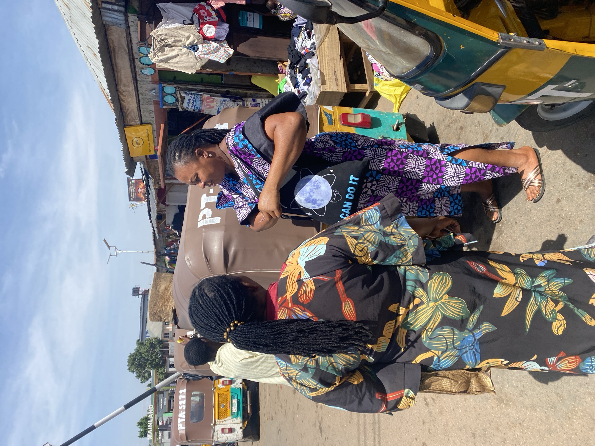 A street scene in Gura Top, Nigeria, shows two women beside parked tricycle taxis. One woman is wearing a purple and blue patterned dress and a black bag with a globe graphic and the words "I CAN DO IT." She is looking into a parked tricycle taxi. Another woman wearing a black floral dress and a hairstyle of dreadlocks adorned with gold beads stands beside the tricycle taxi. In the background, there's a store with goods displayed, a yellow MTN (mobile telephone network) sign, and other street details. The sky is visible and partially overcast. A sign reading "Mainland Lubricant Bay, PTA-5171" is also visible.