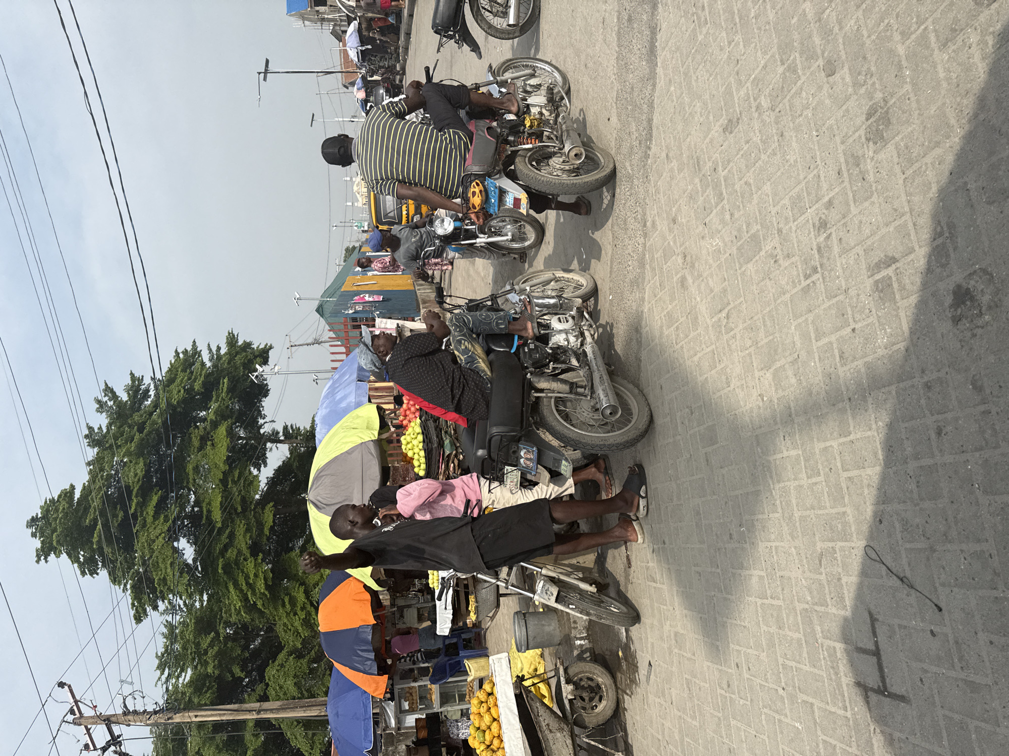 This image depicts a bustling street scene, likely a marketplace or thoroughfare in Ado, Nigeria. The setting is an outdoor area with a ground paved with grey interlocking concrete blocks. The sky is overcast or hazy, indicating daytime with diffused sunlight.
Numerous people are visible throughout the scene, engaged in various activities. Several individuals are associated with motorcycles, commonly known as 'okadas'. In the mid-ground, a man wearing a black and yellow striped shirt is seated on a motorcycle, facing away from the camera, while another man in a grey shirt sits on a motorcycle in front of him. A third man in a black t-shirt and shorts stands near a market stall, his arm slightly raised, looking towards the left. Many individuals in the image are barefoot or wearing simple sandals.
The presence of market activity is prominent, with several makeshift stalls featuring colorful umbrellas (orange, blue, yellow, green) providing shade. One stall prominently displays piles of bright yellow fruits, possibly mangoes or oranges, while another shows red and green produce. A yellow tricycle (known as 'Keke Napep') is partially visible in the background, alongside other parked motorcycles. Overhead, utility poles with a network of electrical wires crisscross the sky, and dense green foliage from trees can be seen in the background, hinting at nearby vegetation. The overall atmosphere suggests a typical day of commerce and transportation in a vibrant community. No clearly decipherable text is visible on any objects.