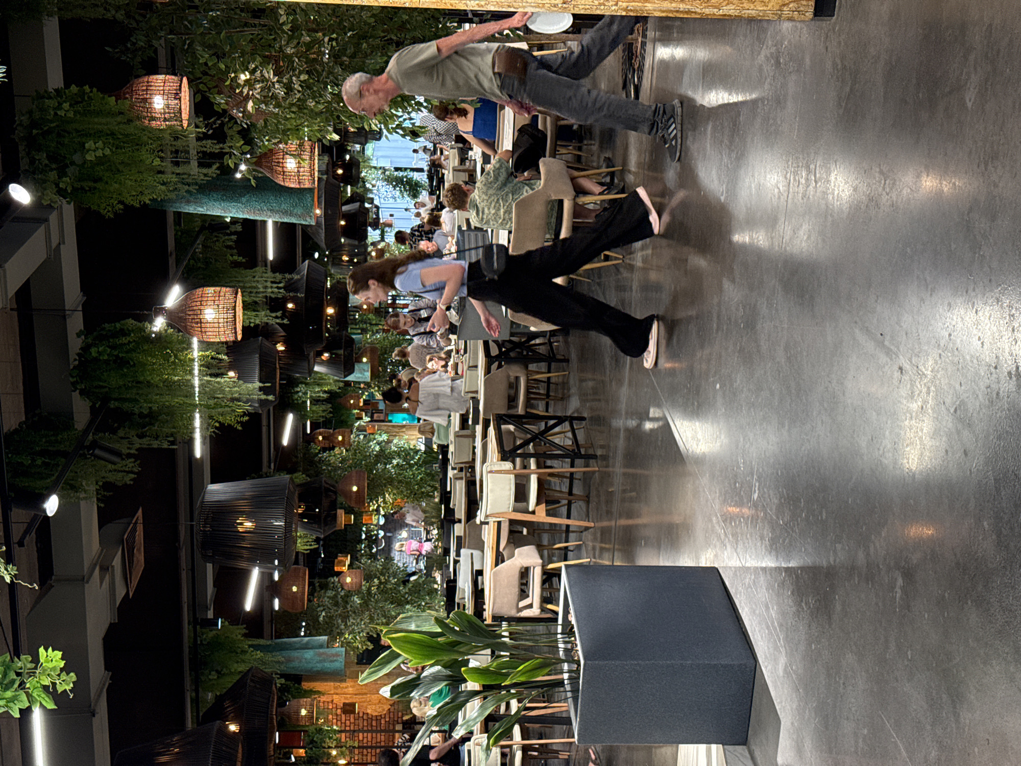 The image depicts the interior of a modern, well-lit restaurant with a strong emphasis on biophilic design, featuring an abundance of hanging plants and lush greenery. The space appears spacious, with a high ceiling from which numerous decorative lamps are suspended, contributing to the ambient lighting. The floor is a polished concrete surface, reflecting the overhead lights and creating a sleek, contemporary aesthetic.
In the foreground, a tall, dark gray planter holds a vibrant green plant with large, elongated leaves. To the right of this planter, a woman with long, dark hair is walking from left to right, wearing a light blue t-shirt and flowing black pants. She is carrying a dark-colored purse on her right shoulder. Further to the right, a man with short gray hair and glasses is also walking from left to right. He is wearing a light green t-shirt, dark gray pants, and athletic shoes, and is holding a white plate in his right hand.
The middle ground of the image shows a seating area with multiple tables and chairs. The tables are made of wood with dark frames, and the chairs are upholstered in neutral tones. Several patrons are seated at these tables, engaged in dining and conversation. In the background, more seating arrangements and decorative elements are visible, including exposed brick walls and further greenery. The overall impression is of a lively and stylish restaurant environment.
The location is identified as the city of Yalta, Ukraine.