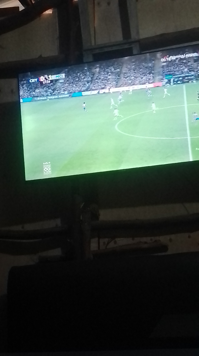 A television displays a soccer game between CRY and MCI. The score is CRY 0-1 MCI at the 57:59 mark. The game is likely a Premier League match based on the on-screen graphics. Players are visible on the field in black and white uniforms. Seating is visible on the edges of the field.