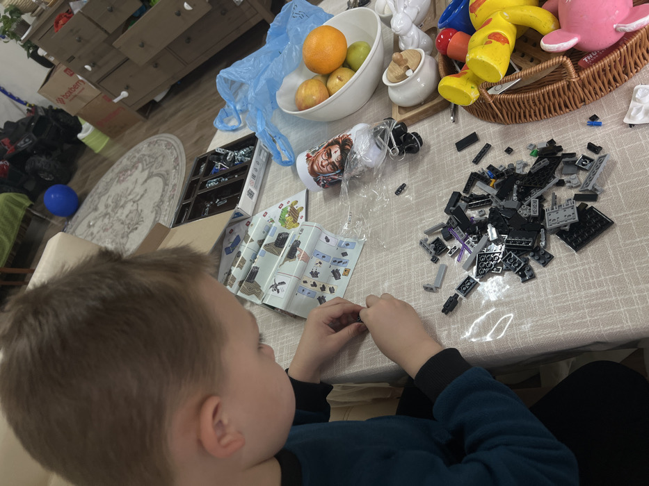 The scene depicts an indoor domestic environment, identified as being in the city of Leninakan, Russia. A young individual with short brown hair, wearing a dark teal or navy long-sleeved shirt with black cuffs, is positioned at a table. The individual is viewed from the top-back of the head and left profile, focused downwards. Their hands are engaged in assembling small black Lego bricks.
The table is covered with a light-colored, possibly patterned tablecloth. Spread before the individual are open instruction booklets for a Lego building set, along with a prominent pile of assorted black and grey Lego pieces. Scattered among these are a few small purple and green bricks.
To the right of the Lego assembly area, a white travel mug featuring a graphic print (possibly of a character) lies on its side, partially encased in clear plastic. Adjacent to this is a crumpled blue plastic bag. A white bowl contains an assortment of fruit: one orange, two red apples, and one green apple. Further to the right, a small white ceramic container with a wooden lid is visible. In the far right foreground, a brown woven wicker basket holds various children's toys, including yellow and blue plastic items and a pink plush bunny.
The background reveals a carpeted floor with an oval, ornate patterned rug. A blue balloon rests on the floor to the left of the rug. A dark-colored ride-on toy, resembling an ATV, is positioned further back. On the right side of the background, a light-colored wooden chest of drawers or cabinet is visible, with at least one drawer partially open. A brown cardboard box bearing the "Boxberry" logo is situated behind this furniture. A light-colored sofa or armchair is partially visible in the left background. The room is illuminated by ambient indoor lighting.