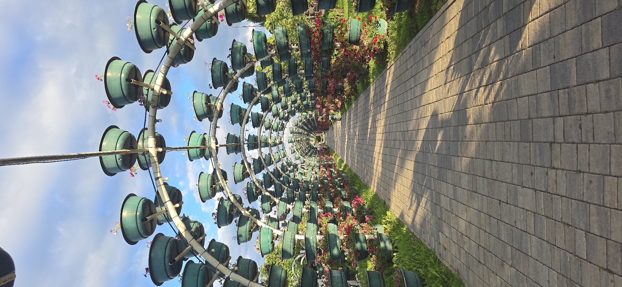 The image depicts a flower tunnel located in Grozny, Russia. A paved pathway with square pavers runs through the tunnel. Arched metal structures support rows of green flower pots containing plants with pink and white flowers. The structures create a tunnel effect, receding into the distance. Sunlight casts shadows on the pathway. The sky is visible in the background, featuring a light blue gradient with some clouds. Green grass and additional flowering plants line the sides of the pathway. No people are visible in the image.