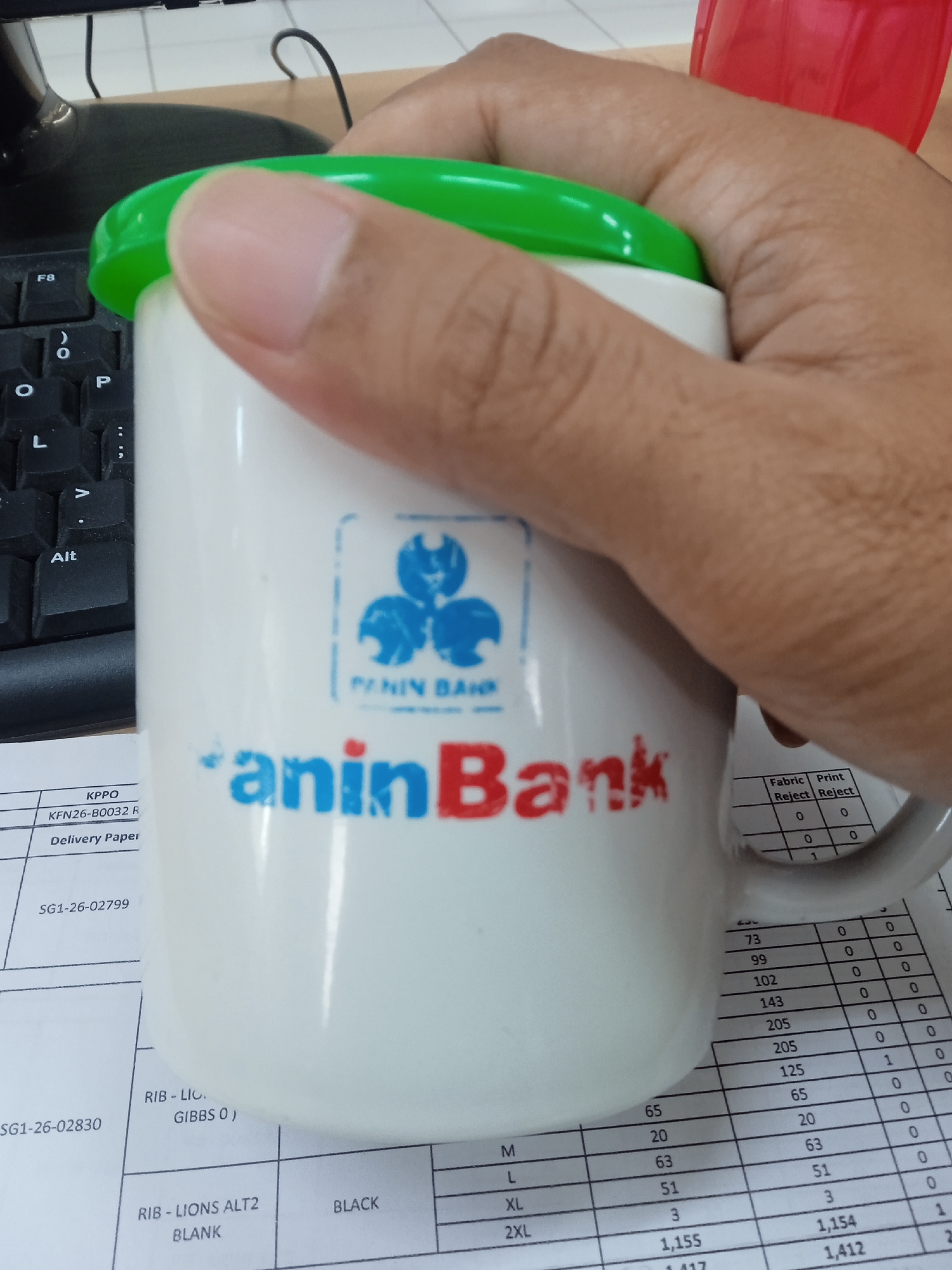A person's hand is holding a white mug with a green lid. The mug has a blue logo with three intertwined shapes and text that reads "PANIN BANK" below it. The words "aninBank" are printed in large blue and red letters on the mug. The mug is placed over a document with a grid of numbers and text. In the background, a black keyboard is partially visible, as is a red plastic cup. The scene appears to be indoors, possibly in an office setting.