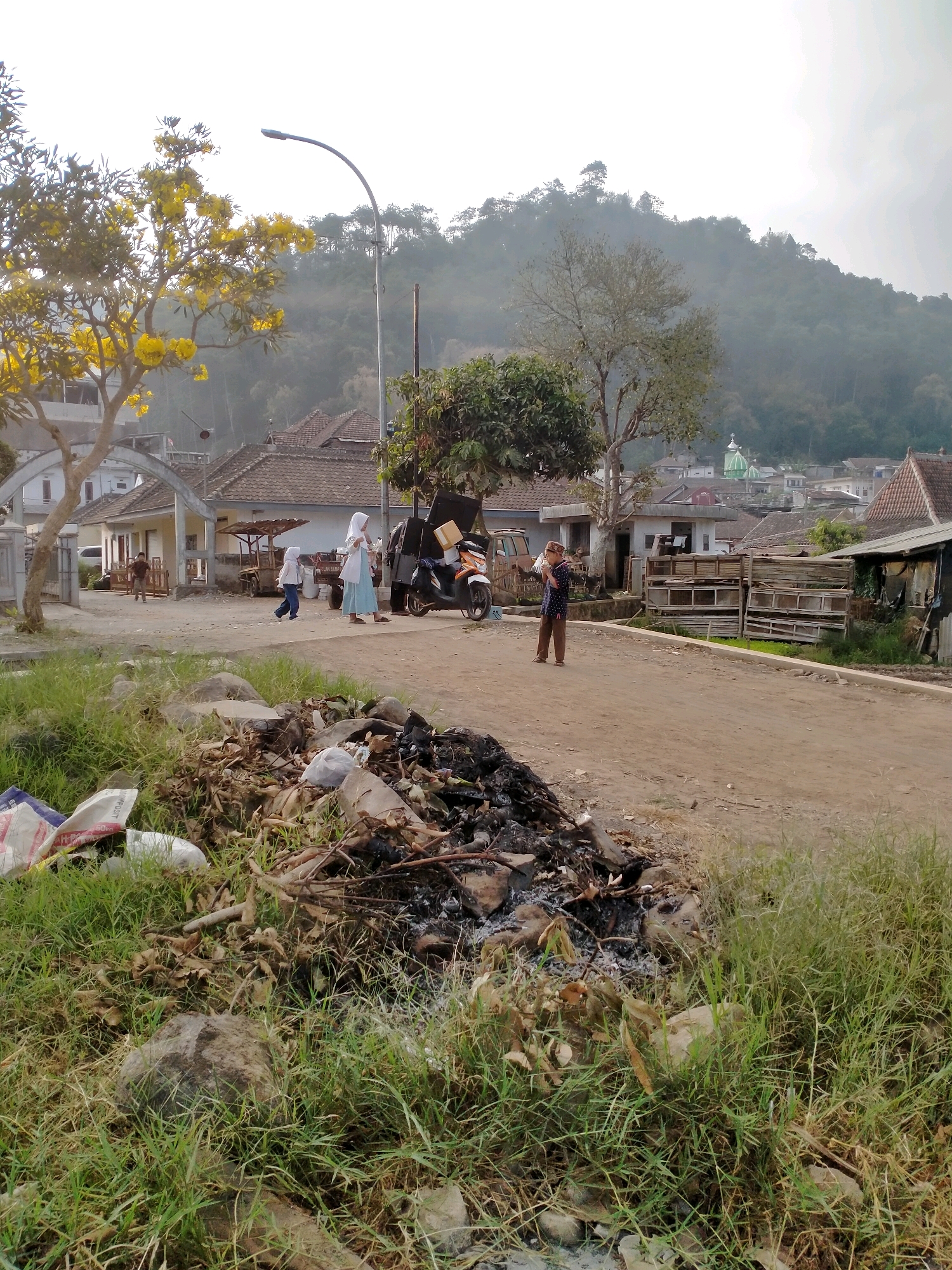 The image depicts a scene in the city of Batu, Indonesia, focusing on a dirt road in a rural or semi-rural area. In the foreground, a pile of garbage and burnt debris is situated on the grassy verge. Beyond this, the dirt road stretches into the middle ground. Several people are present. Two young individuals, possibly children, are walking on the road, one wearing a white headscarf and the other a blue top and white pants. An adult wearing a white headscarf and a blue dress stands nearby. A man in a patterned shirt and brown pants is seen holding something and looking towards the right. A motorcycle with a large box attached to the back is parked on the road. In the background, houses with tiled roofs are visible, typical of Indonesian architecture. A prominent street lamp stands to the left of center. Further in the distance, a lush green hill or mountain rises, partially shrouded in mist or haze, with a mosque or similar building with a green dome visible on its slopes. The overall atmosphere suggests an outdoor, daytime setting with a somewhat overcast sky.