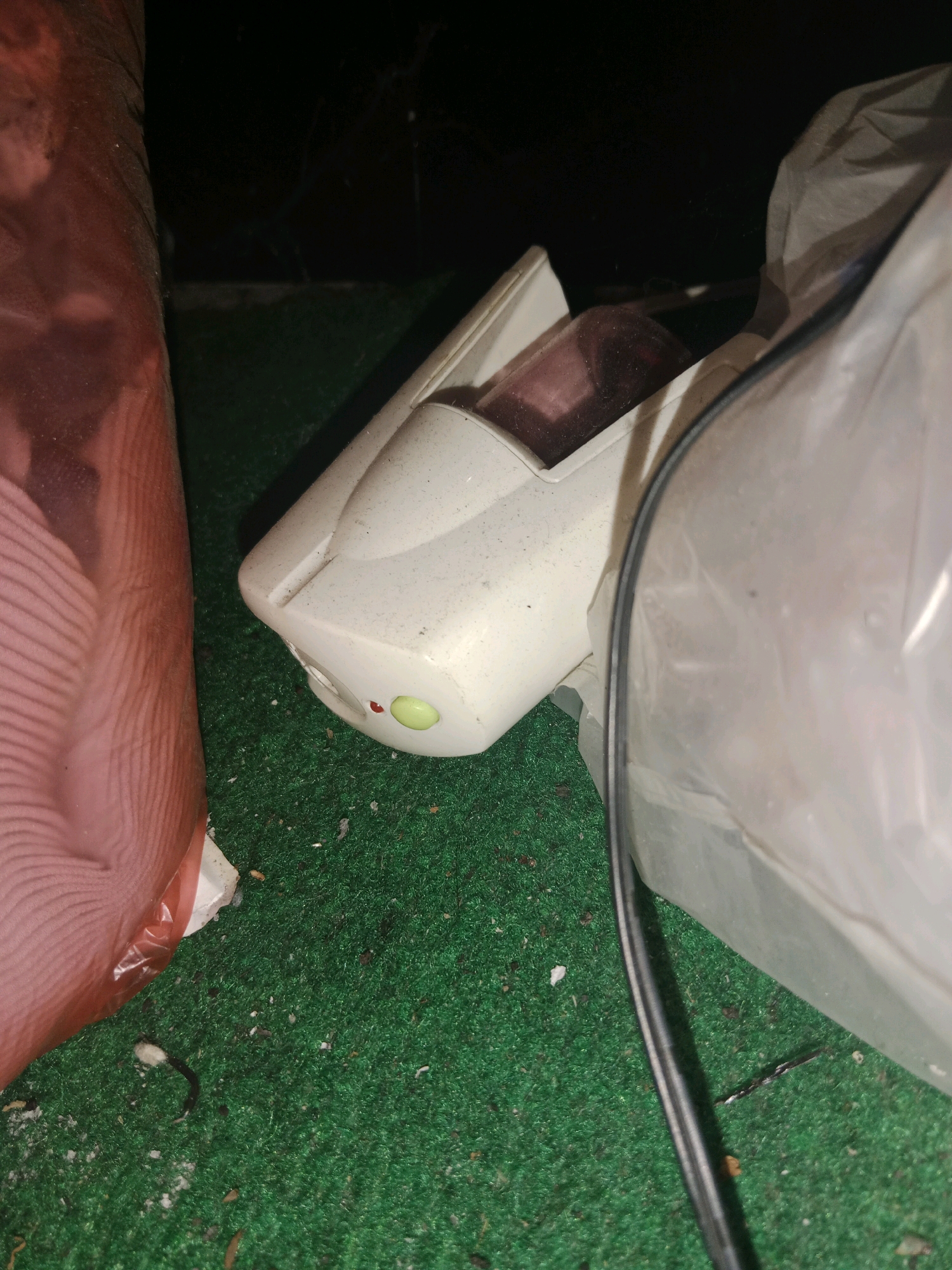 The image shows a close-up, low-angle view of a white, futuristic-looking device, possibly a remote control or a small appliance, resting on a green, carpet-like surface. The device has a transparent red-tinted window on top and a green button or light on its front. To the left, a large, translucent pink object with visible vertical ridges, wrapped in plastic, takes up a significant portion of the frame. To the right, a crumpled, translucent white plastic bag partially obscures another part of the device and the background. A black cable runs diagonally across the foreground, disappearing behind the white plastic bag. The overall lighting is dim, suggesting an indoor or nighttime setting. The surface appears to be artificial turf, possibly in a residential or storage area. There is no indication of the specific location within Ringinrejo, Indonesia, beyond the subject matter presented in the image.