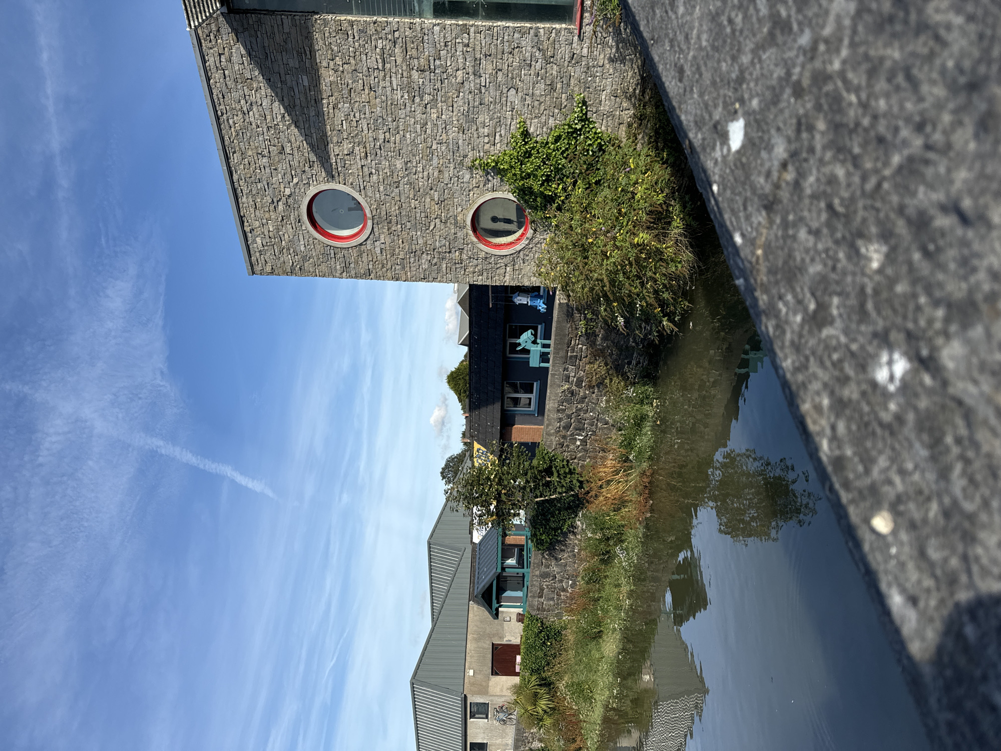 The image depicts a riverside scene in Waterford, Ireland. In the foreground, a stone wall runs along the edge of a body of water. The water reflects the sky and the buildings on the opposite bank.
The most prominent building is a modern structure with a stone facade and two circular windows with red trim. To the left of this building, a row of older structures runs along the riverbank. These include a building with a corrugated metal roof, another with a dark roof and a turquoise structure in front of it, and a building with a dark roof and a light-colored wall.
There is vegetation along the riverbank, including bushes and some taller trees. The sky is blue with wispy clouds. A faint contrail from an aircraft is visible in the upper left portion of the sky. The overall scene suggests a calm, daytime setting.