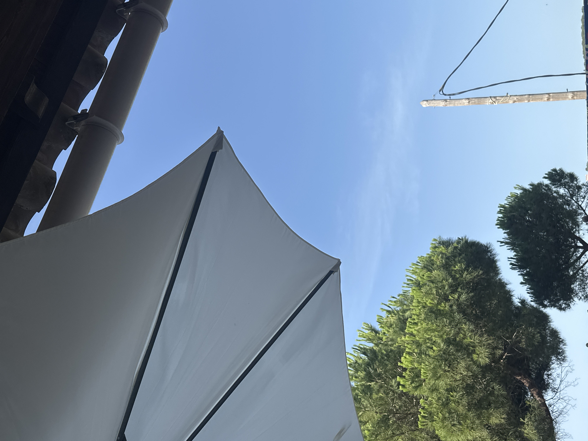 The image captures a bright, sunny day in Sant Feliu de Guíxols, Spain. The foreground is dominated by a large, white umbrella tilted to the left, its fabric taut and casting a shadow. Beneath the umbrella, lush green pine trees are visible, their needles dense and vibrant against the clear blue sky. In the upper right quadrant, a section of a building's roof structure with dark wooden beams and a beige downspout is attached to the eaves. A weathered wooden pole with electrical wires strung between it and an unseen point stands to the right, also set against the expansive blue sky. The overall scene suggests an outdoor setting, possibly a patio or garden area, enjoying a clear day.