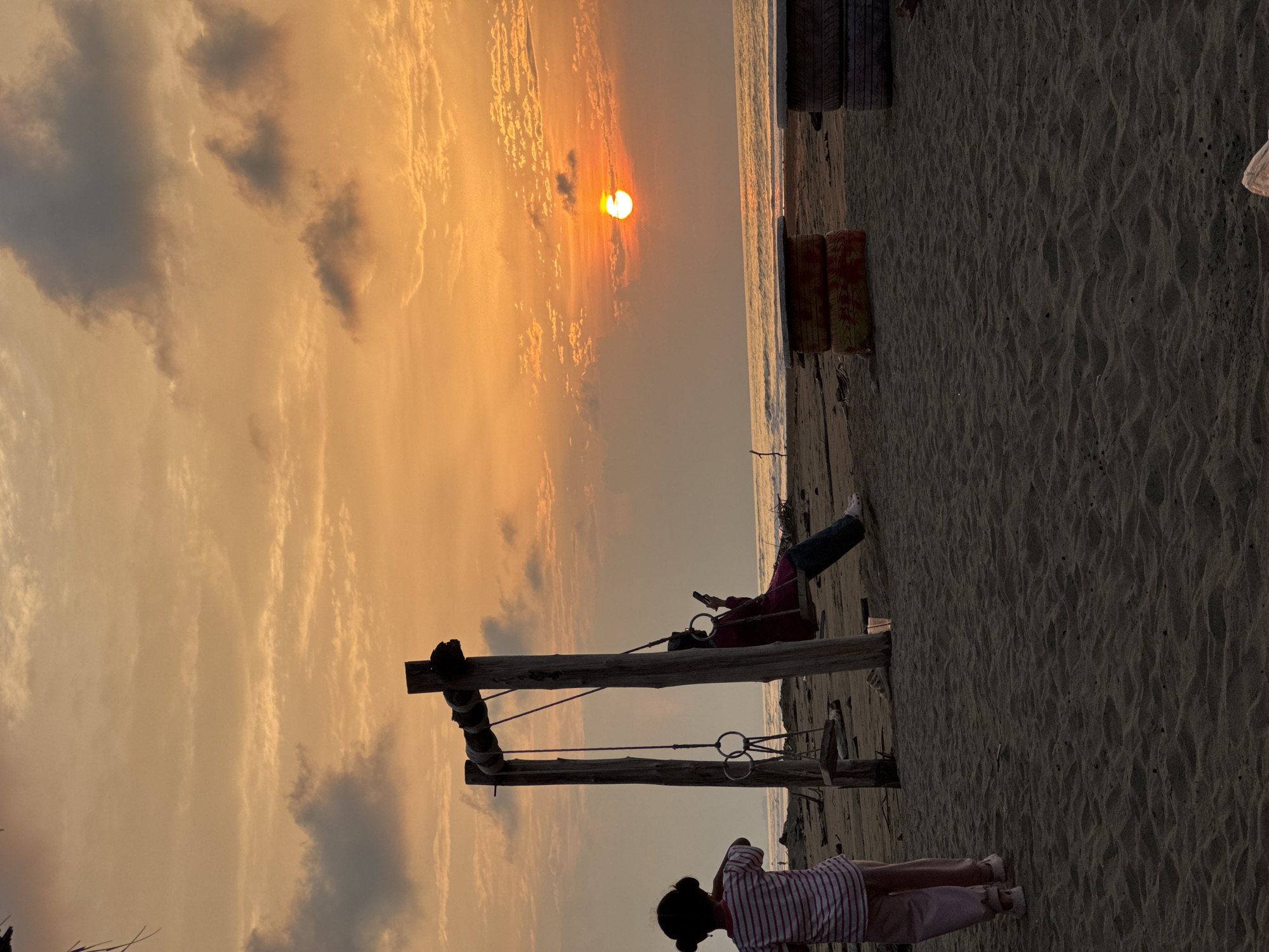 The image captures a serene beach scene at sunset in Reudeuep, Indonesia. The sky is ablaze with warm orange and yellow hues, with scattered clouds adding texture. The sun, a bright orb, is setting over the calm ocean, casting a reflection on the water's surface.
In the foreground, a sandy beach stretches out. A rustic wooden swing set stands near the center, with two vertical posts and a horizontal beam from which a swing is suspended. One person, partially obscured, is seated on the swing, looking at their phone. To the left of the swing set, a young child with dark hair tied in two buns is standing, facing away from the camera, towards the ocean. The child is wearing a striped long-sleeved shirt and pink pants.
Further back on the beach, two stacks of what appear to be old tires, repurposed as makeshift tables, are visible. Debris and driftwood are scattered across the sand. The overall atmosphere is peaceful and tranquil, highlighting the natural beauty of the coastal landscape during the golden hour.