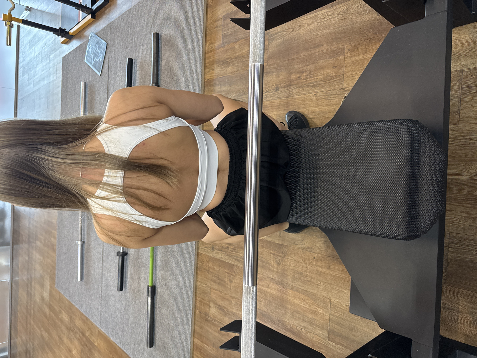 A woman with long brown hair is lying on a black weight bench in what appears to be a gym. She is wearing a white sports bra and black shorts. A barbell is positioned above her. The gym has a wood floor with a gray mat to the left. Various pieces of gym equipment, including weights and a squat rack, are visible. The location, Chaikovsky, Russia, cannot be confirmed from the image.