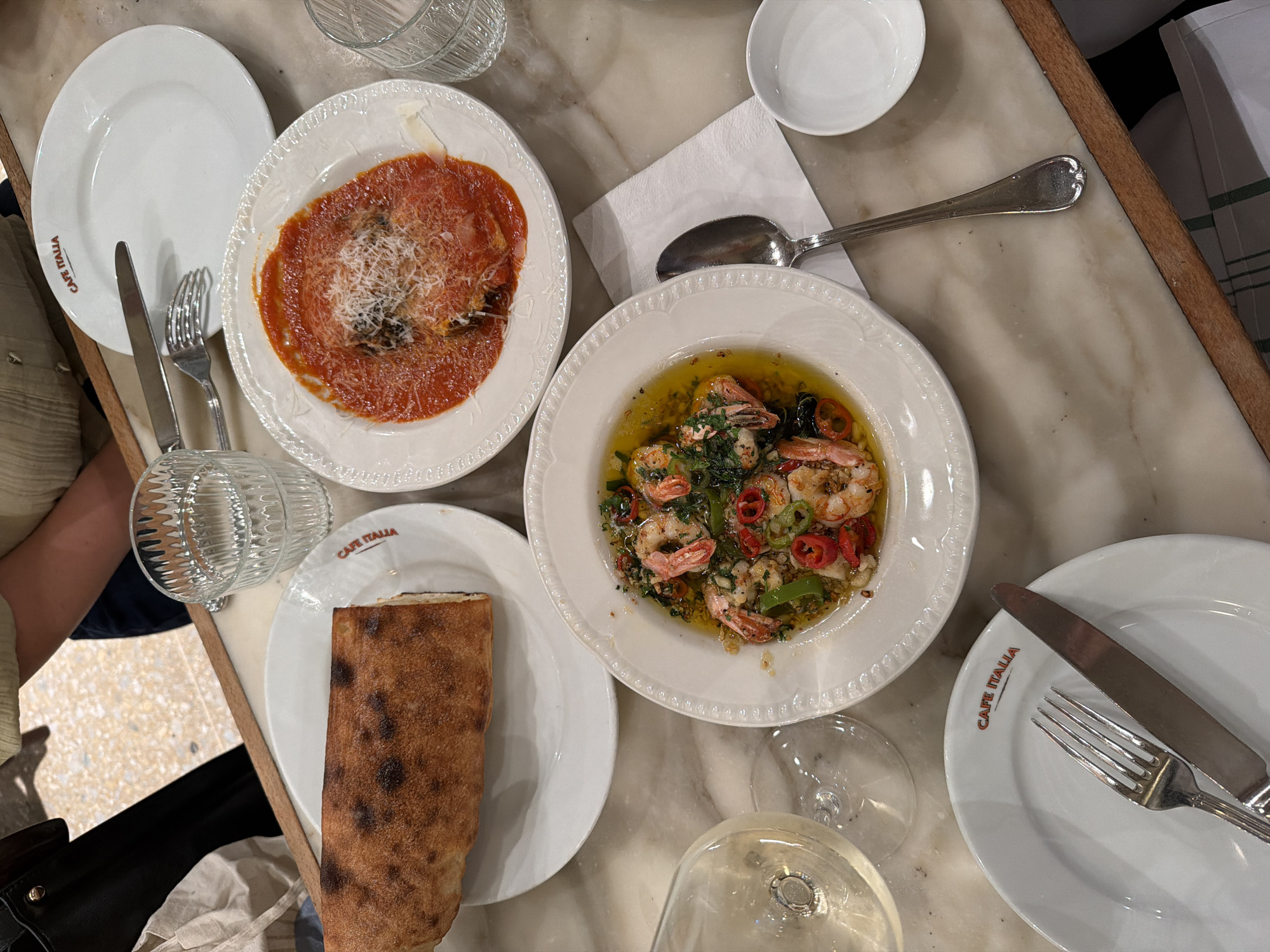 The image shows a table at Cafe Italia, Tel-Aviv, Israel. Several plates are arranged on a marble-patterned table.
One plate contains a dish with red sauce and grated cheese. Another holds shrimp cooked in oil, herbs, and peppers. There is a dish of bread next to the shrimp and a separate empty plate.
Silverware (forks, knives, and a spoon) are also on the table. There are two glasses, one containing liquid. A small white bowl sits near a folded white napkin.
The logo "CAFE ITALIA" is visible on some of the plates. The scene appears to capture a meal in progress. A person is visible on the left side of the frame.