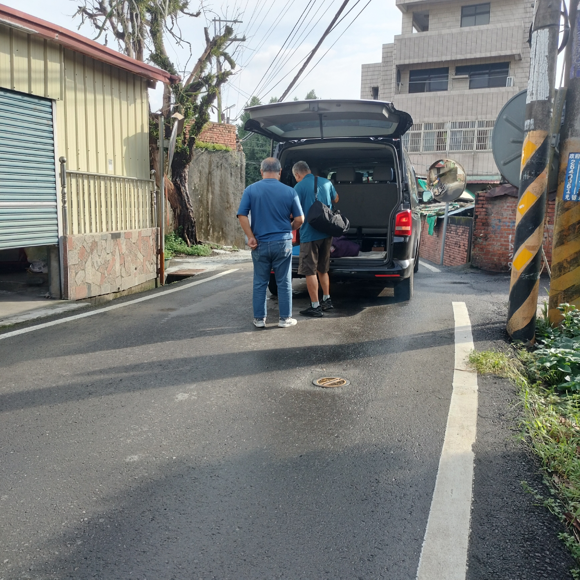 Two men are standing by the open rear door of a black minivan on a paved road in Zhushan Township, Taiwan. The man on the left is wearing a blue t-shirt and blue jeans, with his hands behind his back. The man on the right is wearing a teal t-shirt, brown shorts, and sneakers. He has a black duffel bag slung over his shoulder and appears to be reaching into the back of the minivan. Inside the minivan, luggage is visible. To the left of the road is a light-yellow building with a corrugated metal exterior and a closed roll-up door. A metal fence with vertical posts and horizontal railings stands in front of the building. A large, gnarled tree with sparse foliage is to the right of the building. On the right side of the road, a utility pole with black and yellow diagonal stripes is prominent. Further down the road, a multi-story building with a brick facade is visible. The road has a white line marking the lane. Overcast skies are present.