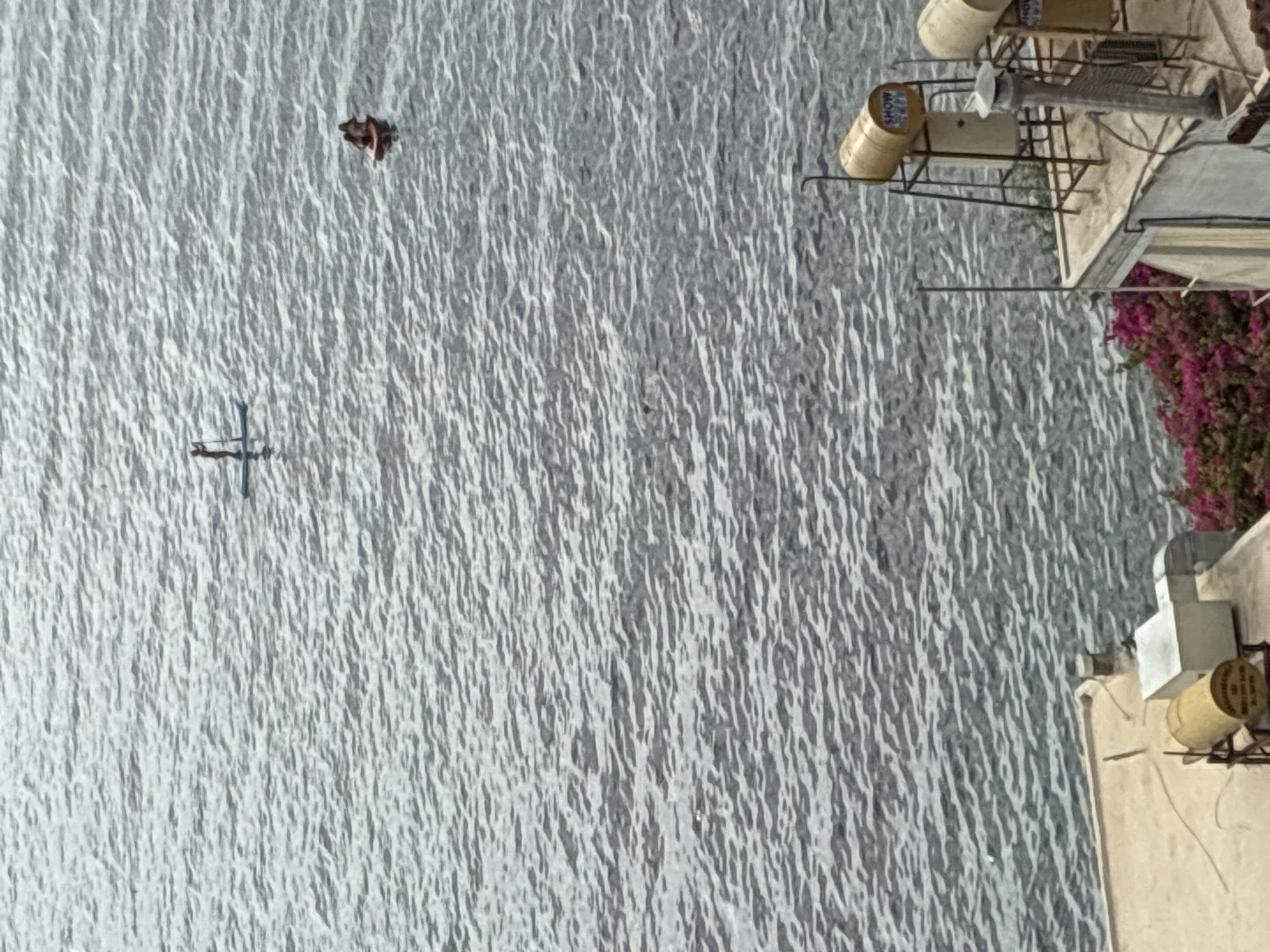 The image shows a body of water, likely the Mediterranean Sea, with two individuals engaged in water activities. In the distance, a person stands on a paddleboard. Closer to the foreground, another small vessel, possibly a dinghy or raft, carries two individuals. The water exhibits a choppy texture with visible ripples. In the lower portion of the image, a rooftop is visible, featuring rooftop water heaters, one of which is cylindrical and yellow with text that reads "SHOW". There is also a white rectangular structure and a metal chimney. A cluster of vibrant pink flowers is present on the lower right side of the rooftop, suggesting a coastal location in Alanya, Turkey, given the presence of such rooftop water systems and abundant flowering plants. The overall scene depicts a leisure activity by the sea with urban elements.