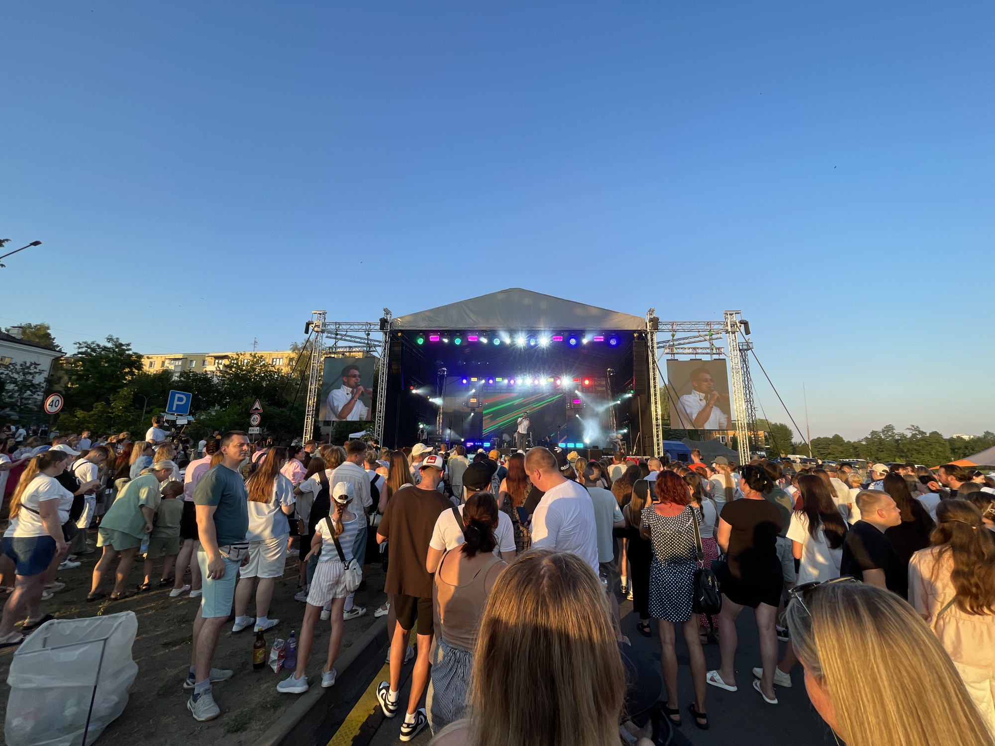 An outdoor concert is taking place in the city of Brest, Belarus. A large crowd of people, dressed in casual summer clothing, are gathered in front of a stage. The stage is equipped with large screens displaying images of performers, and is illuminated by colorful lights. The event appears to be taking place during the late afternoon or early evening, with the sun casting long shadows across the scene. There are trees and buildings visible in the background, suggesting an urban or suburban location. Several signs, including traffic signs and a parking sign, are also present, indicating the presence of public infrastructure. A large trash bag is visible in the foreground, suggesting that the event has just concluded or is ongoing with waste management in place.