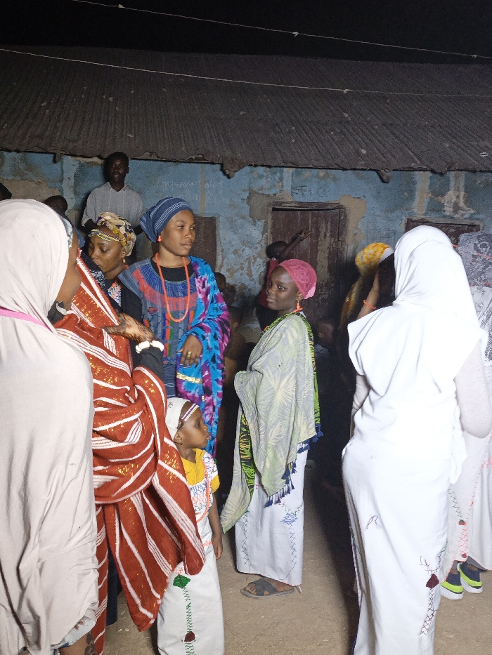 A group of people are gathered outside a building with a corrugated metal roof. Several women are wearing colorful headscarves and wraps, while others are wearing white hijabs. One young girl stands out with a red and white striped garment. A man stands in the background, partially obscured by the building. The overall atmosphere appears to be lively and communal.