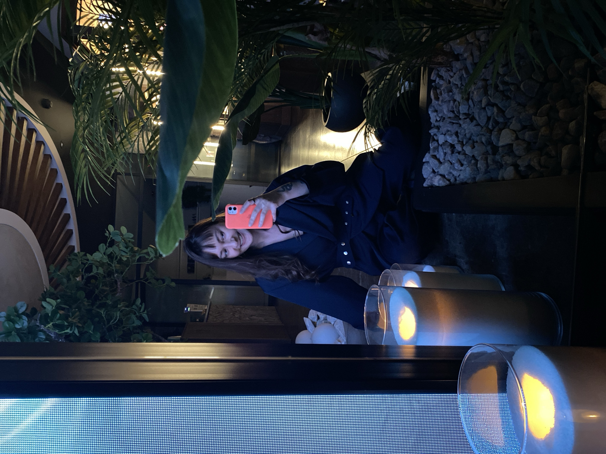 A woman with long brown hair and bangs is holding a pink smartphone and smiling in what appears to be a dimly lit, upscale indoor setting. She is wearing a dark blue or black collared shirt with silver buttons. The background features elements of interior design, including plants, decorative stones, and what appears to be a display of cylindrical glass containers holding candle-like lights. The setting includes architectural elements with wooden accents. The foreground shows a screen with a patterned display. There is no indication of this image being taken in Votkinsk, Russia.