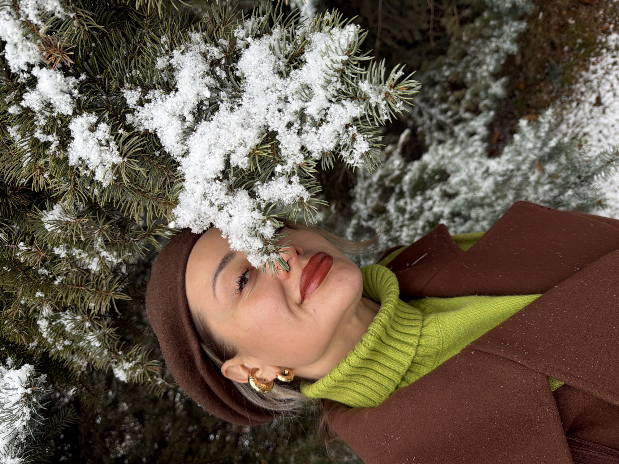 A woman lies in a snow-covered evergreen bush. She wears a brown beret, a chartreuse turtleneck sweater, and a brown coat. She wears gold hoop earrings and red lipstick. The location may be Krasnoyarsk, Russia.