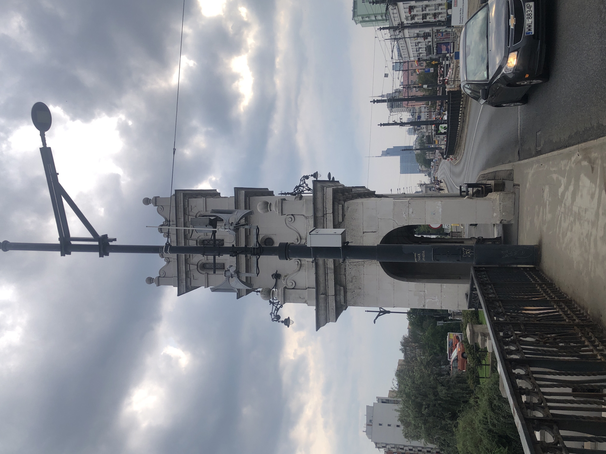 The image depicts a view from a bridge in Warsaw, Poland, looking down a street. In the foreground, a dark gray Chevrolet sedan with license plate WWL 8834P is driving on the road. To the left, there is a pedestrian walkway with a black metal railing. Dominating the center of the image is a large, ornate stone structure, possibly part of the bridge's architecture, with decorative elements and arched openings. A tall, black lamppost with a modern light fixture extends upwards from the structure. In the background, a cityscape with various buildings, including a tall skyscraper, is visible under a cloudy sky. Green trees and foliage line the sides of the street in some areas. The overall scene is urban and appears to be during daytime, with overcast conditions.